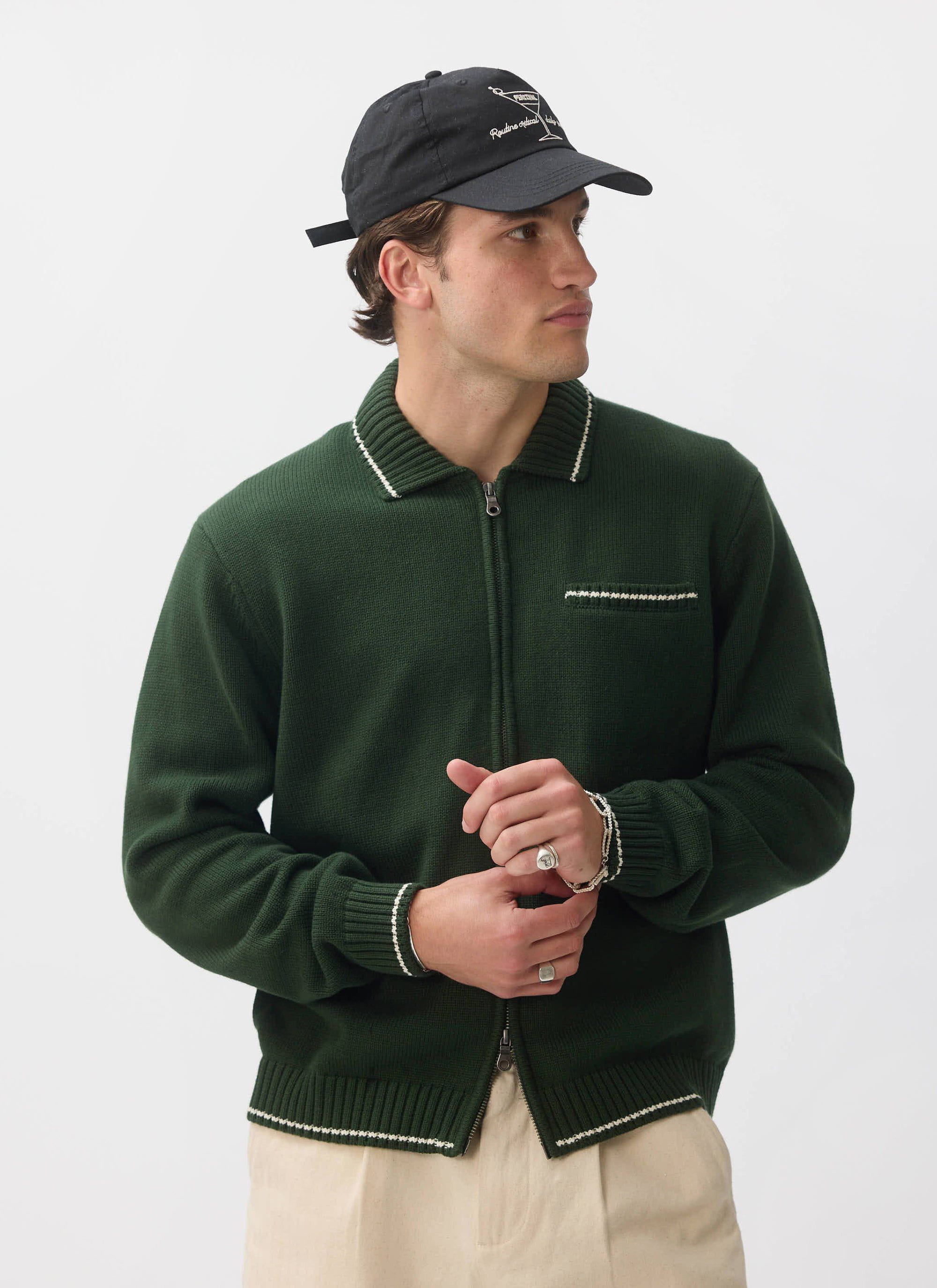 A man models the Percival Gardner Knitted Zip Cardigan in forest green, adjusting his cuff while wearing a black cap.