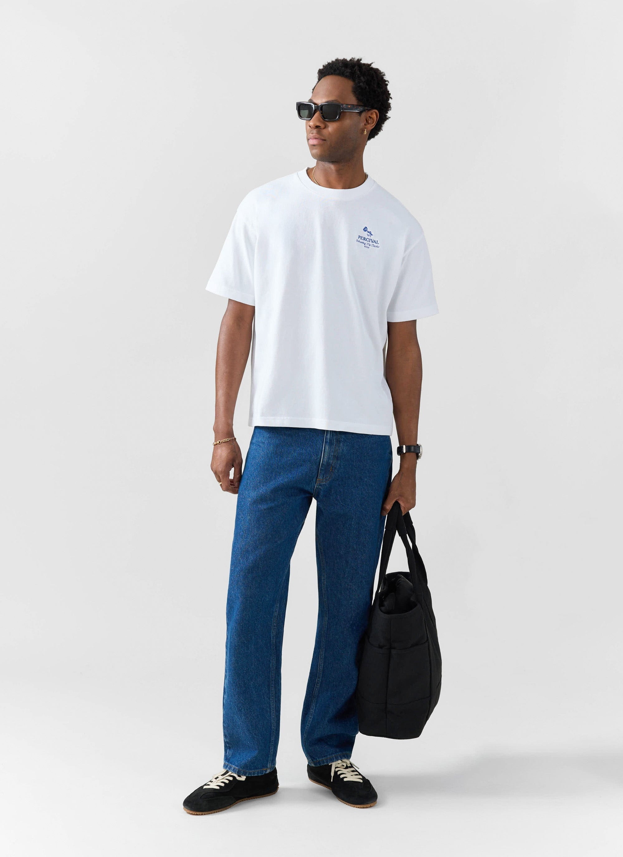 A man models the Percival Smith Floral Oversized T-shirt in white, styled with blue jeans, sunglasses and a black bag.