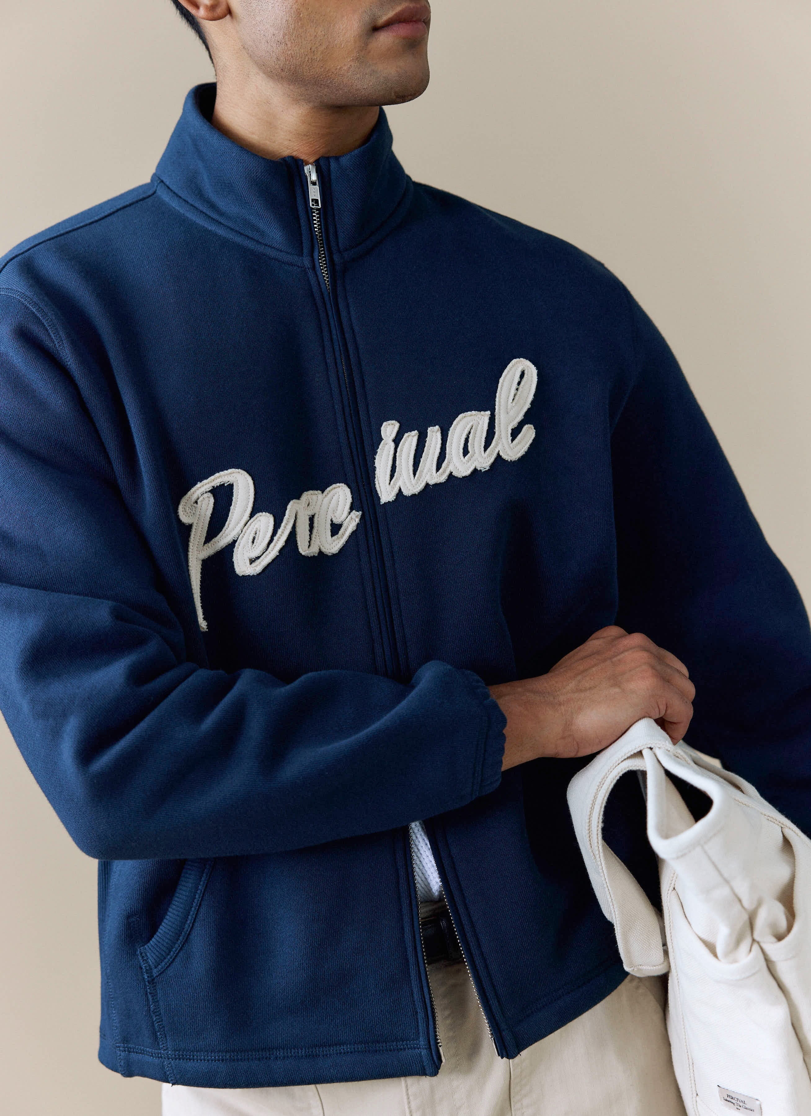 Close up of the white script embroidery on the chest of the navy cotton Curtis Boxy Zip Funnel Neck Harrington from Percival.
