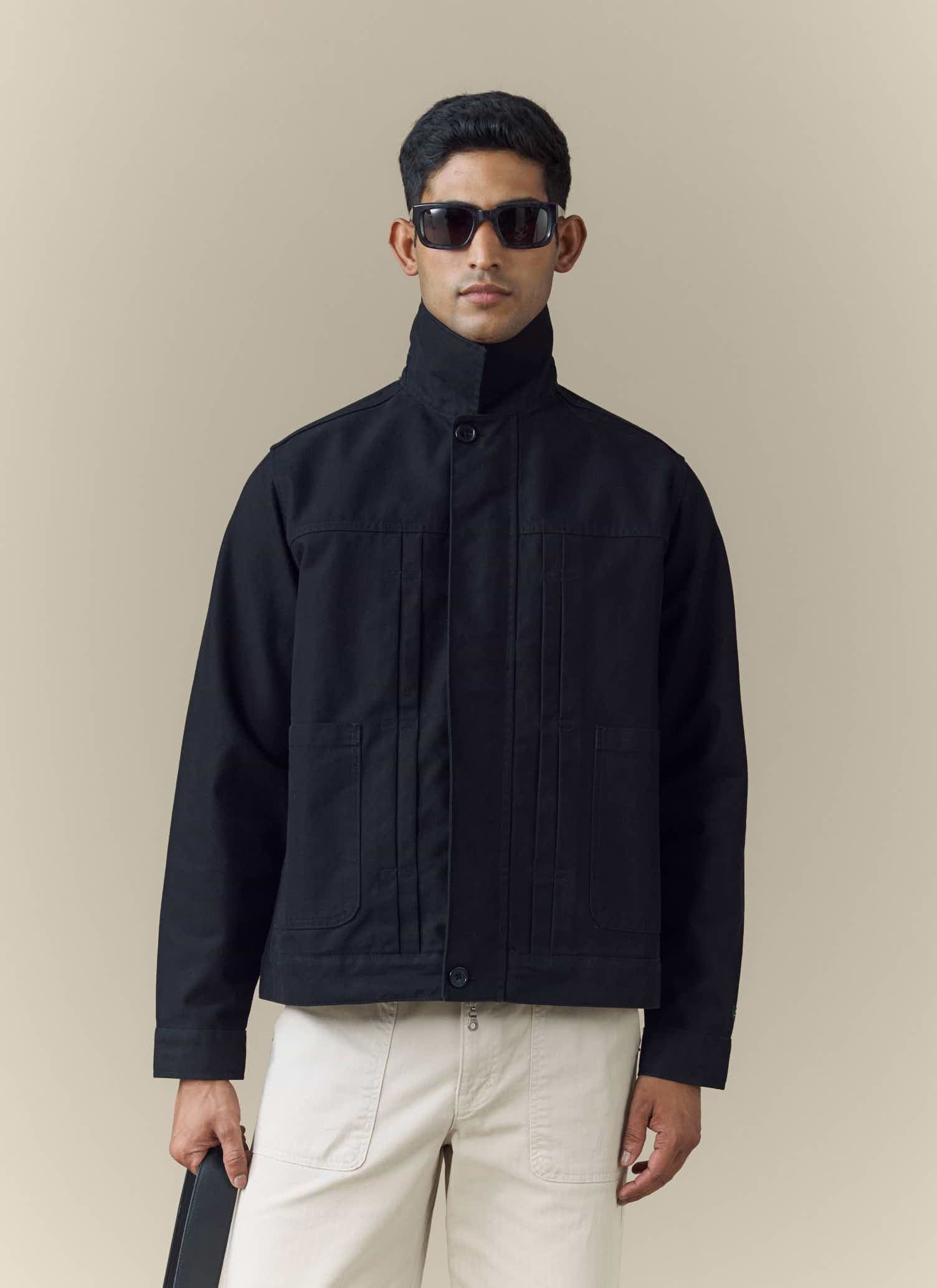 A model wears the black Ernst Jacket from Percival with a high buttoned collar, sunglasses, and light-coloured trousers.