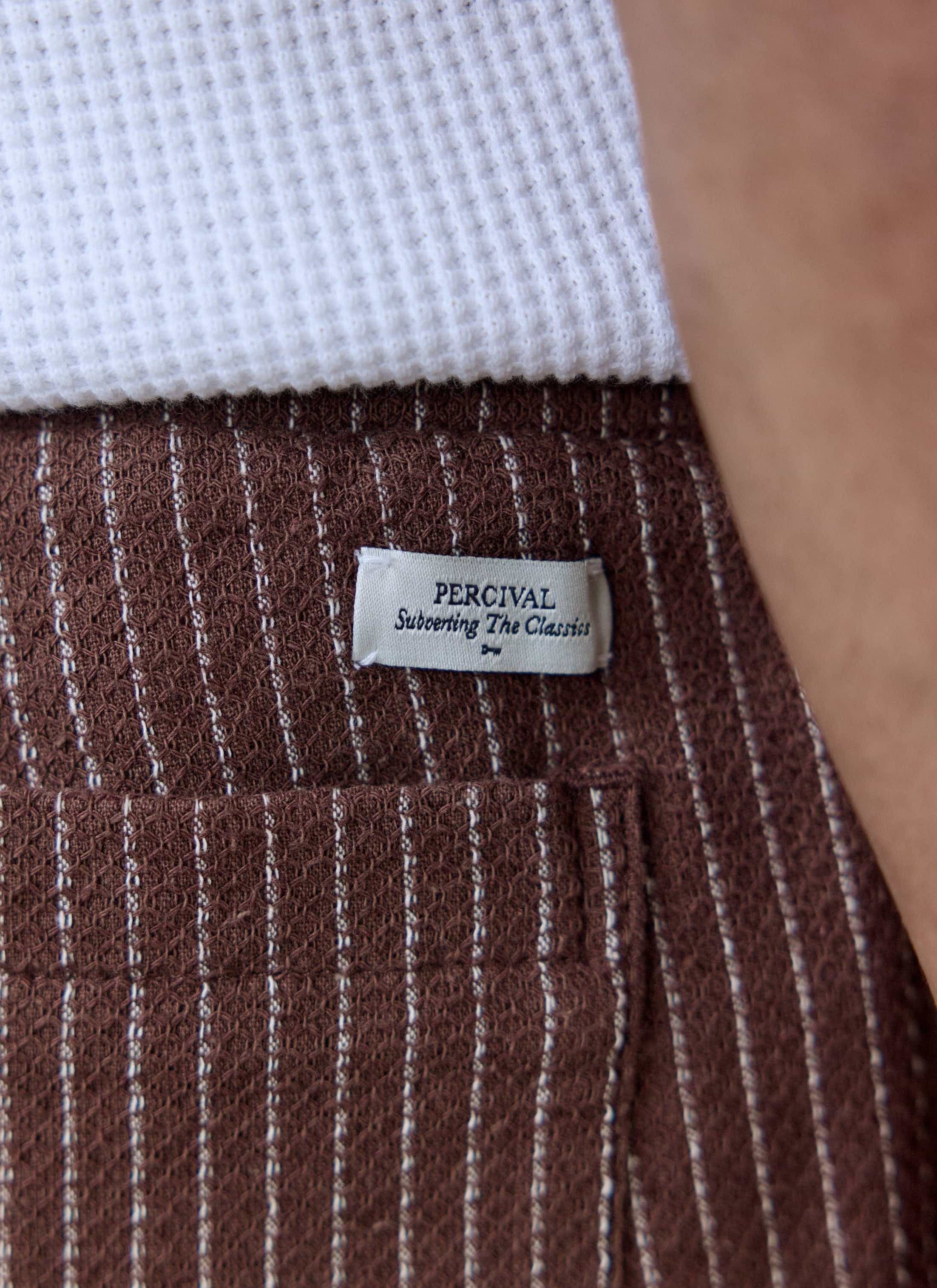 Close-up of the brown linen Sorrento Shorts from Percival, showing the subtle stripe and "Subverting The Classics" label.