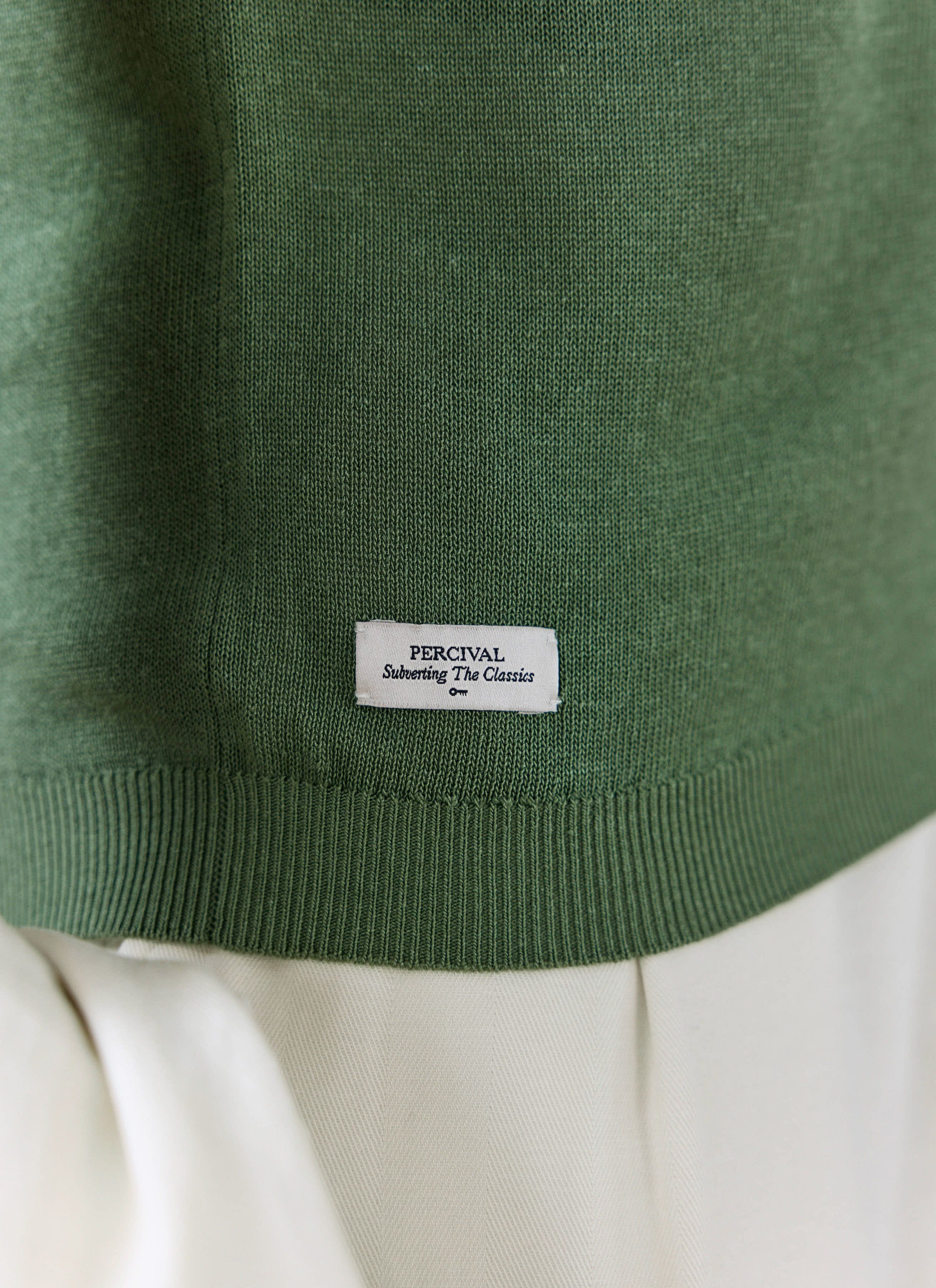 Close-up of the forest green Laurence Knitted Polo by Percival, highlighting the ribbed hem and white logo tag.