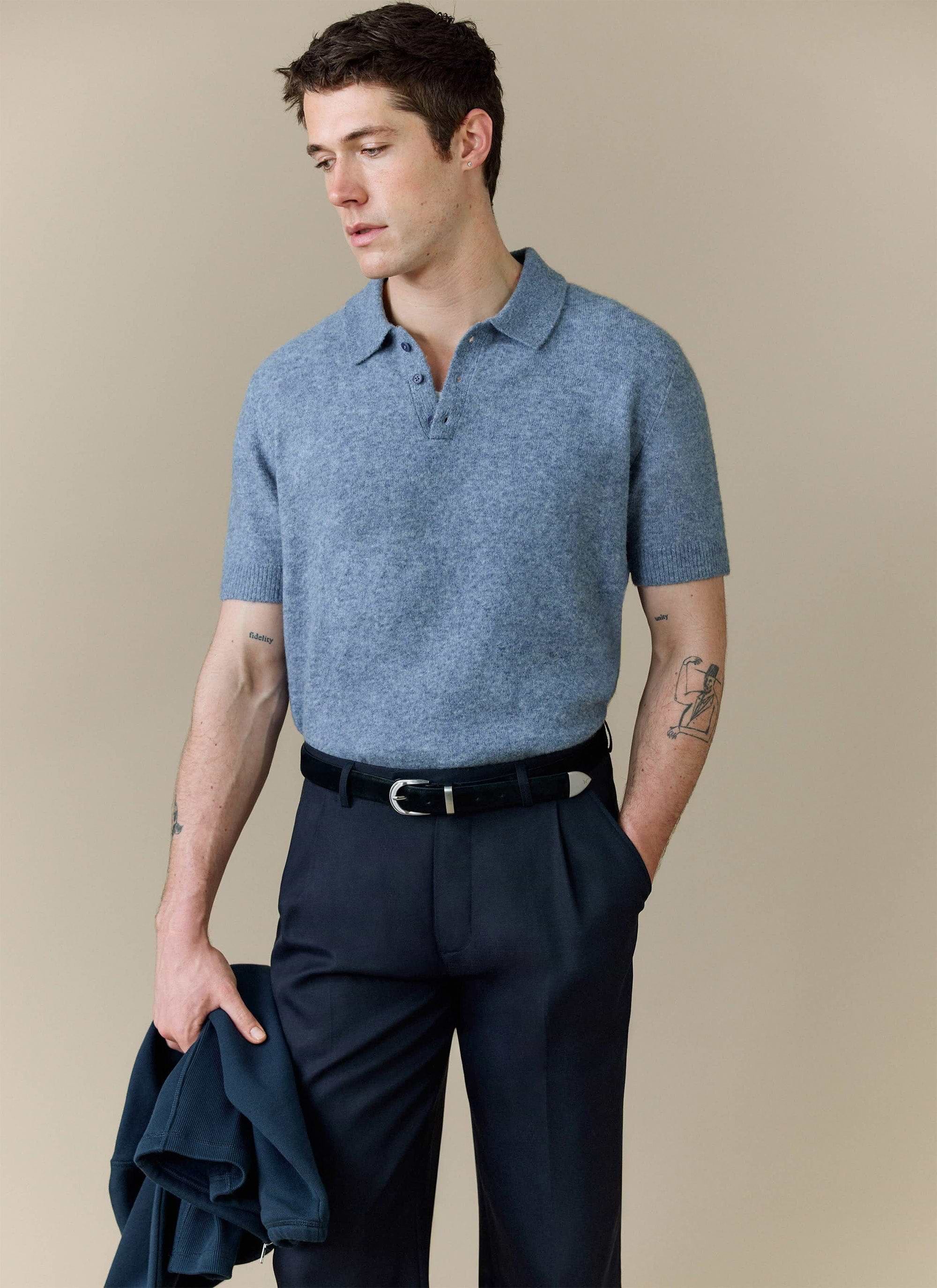 A model wears the Percival Collins Boxy Knitted Polo in blue, styled with smart dark trousers against a studio background.