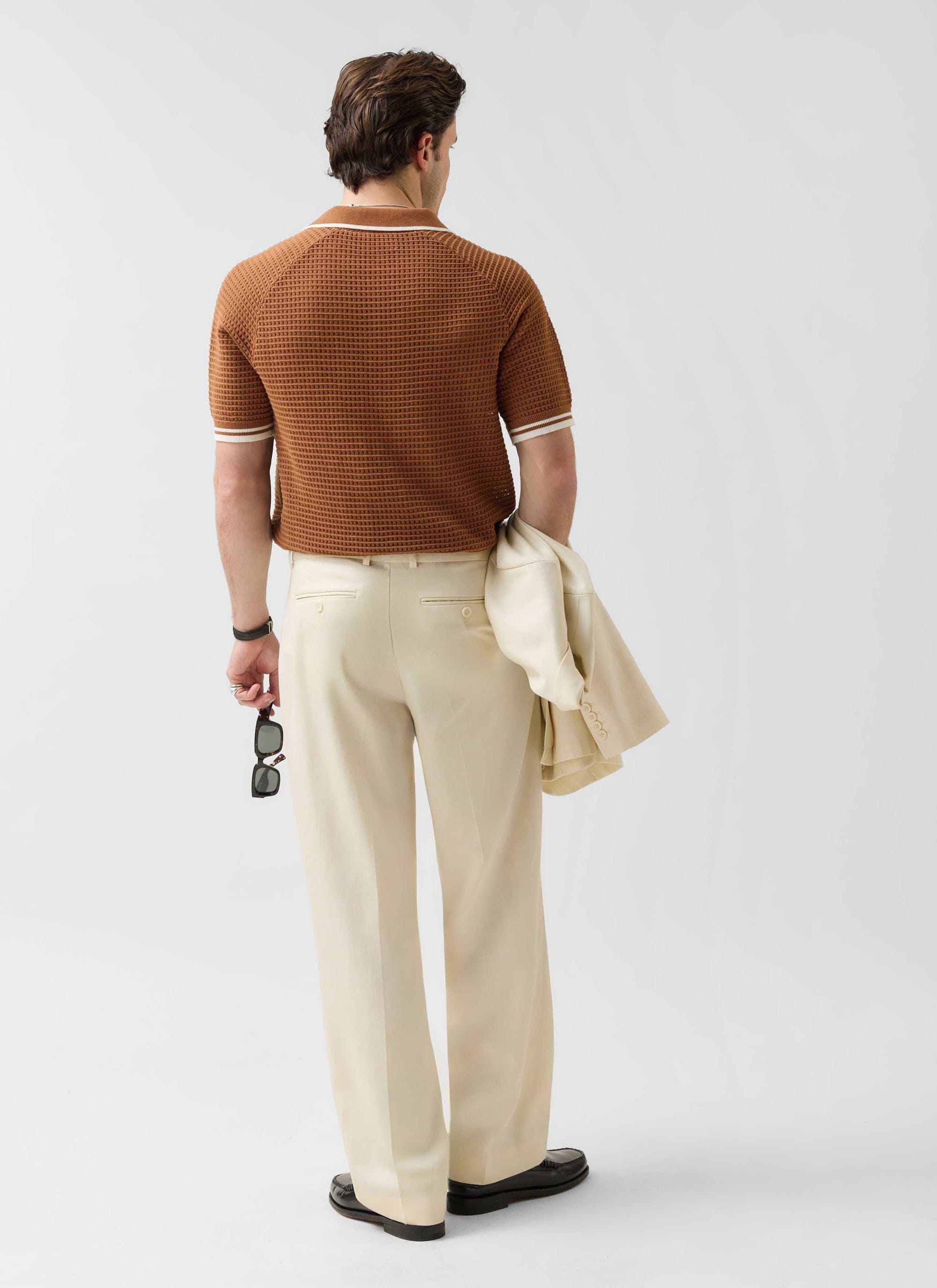 Back view of a model in the Percival Tennis Boxy Knitted Polo in brown, paired with cream trousers and holding a jacket.