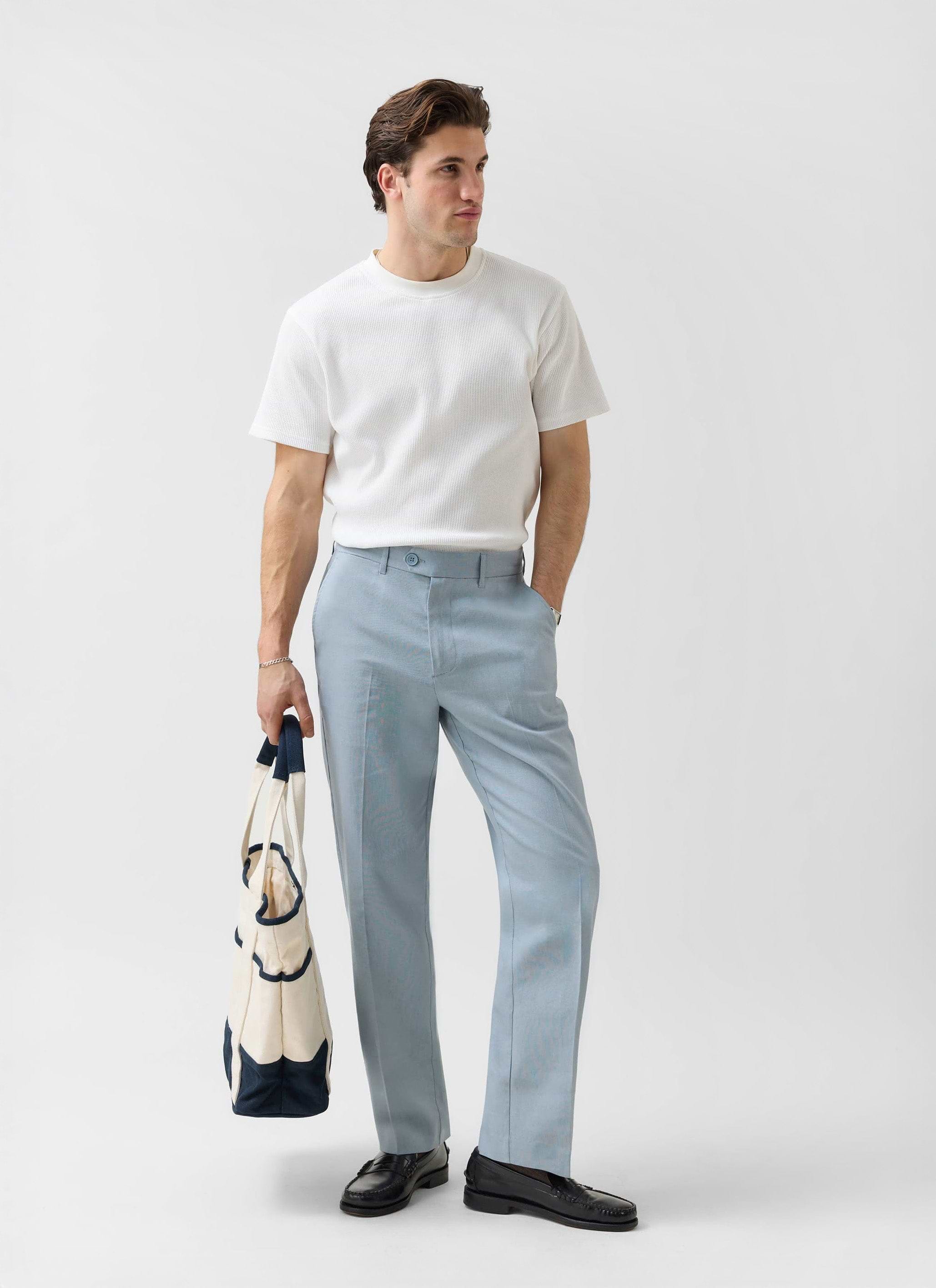 Model wears Percival Menswear light blue tailored linen trousers with a white t-shirt and black loafers, holding a tote bag.