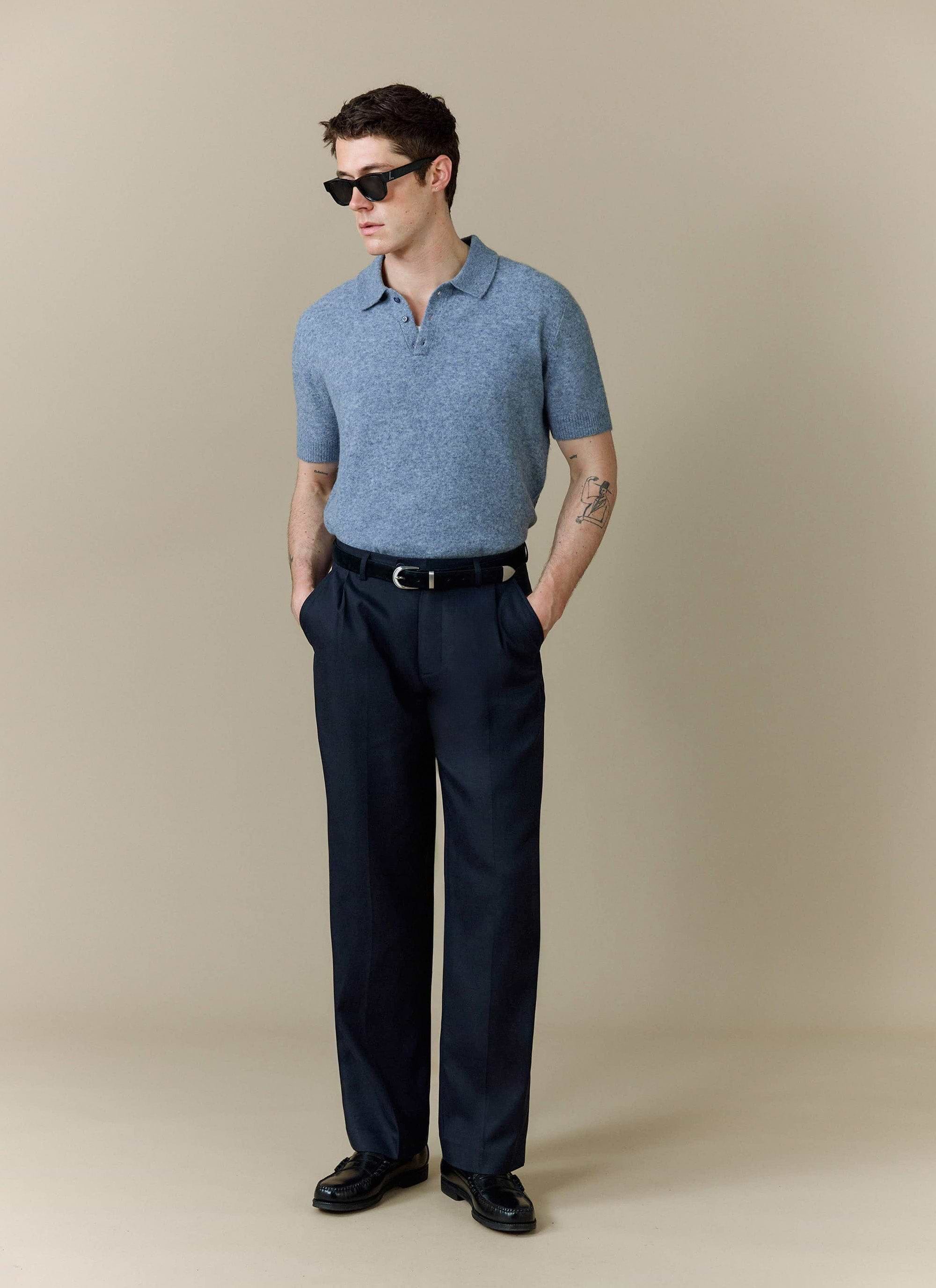 A model wears the Percival Collins Boxy Knitted Polo in blue, styled with smart navy trousers and black sunglasses.