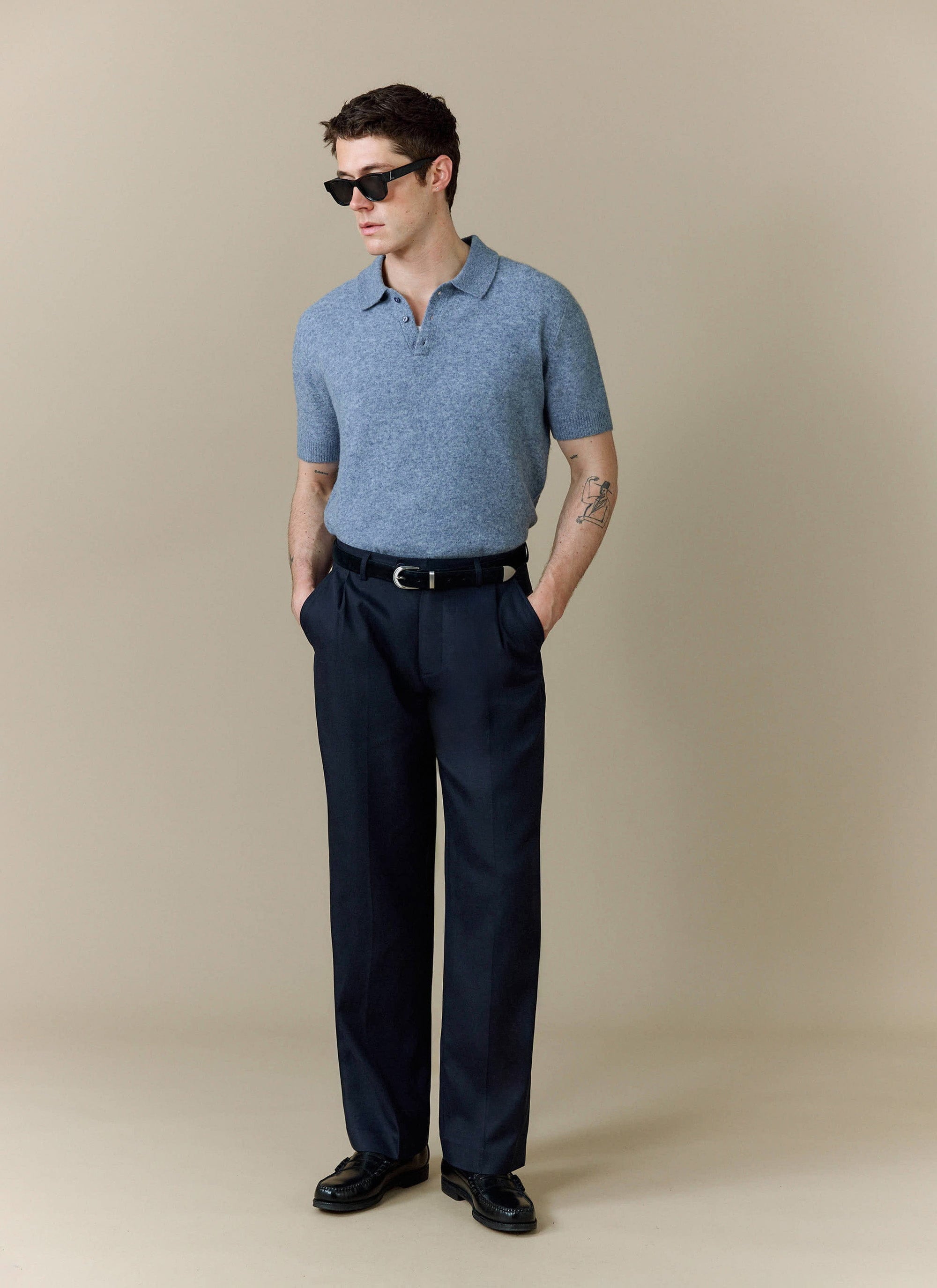 A model wears the Percival Collins Boxy Knitted Polo in blue, styled with smart navy trousers and black sunglasses.
