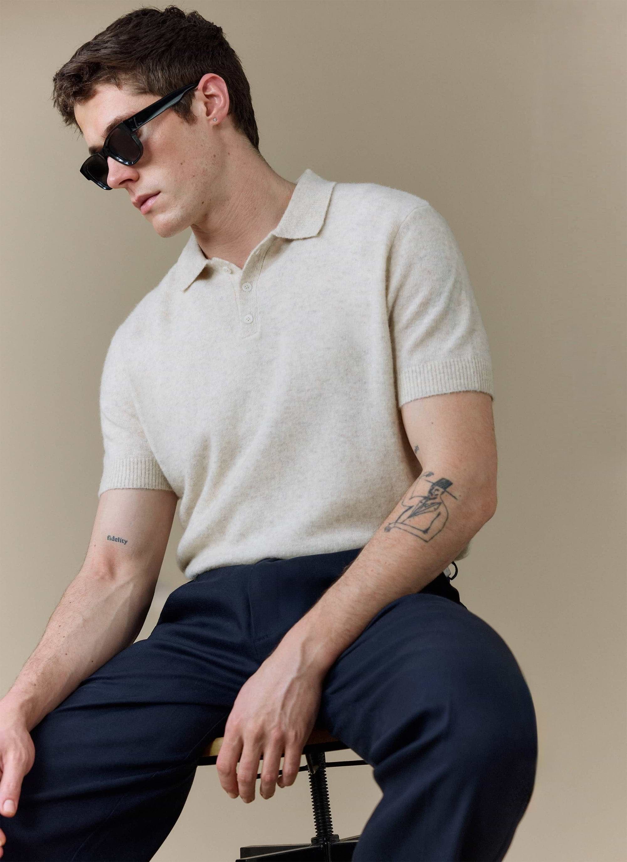 A model sits wearing the Percival Collins Boxy Knitted Polo in ecru, styled with dark trousers and sunglasses.