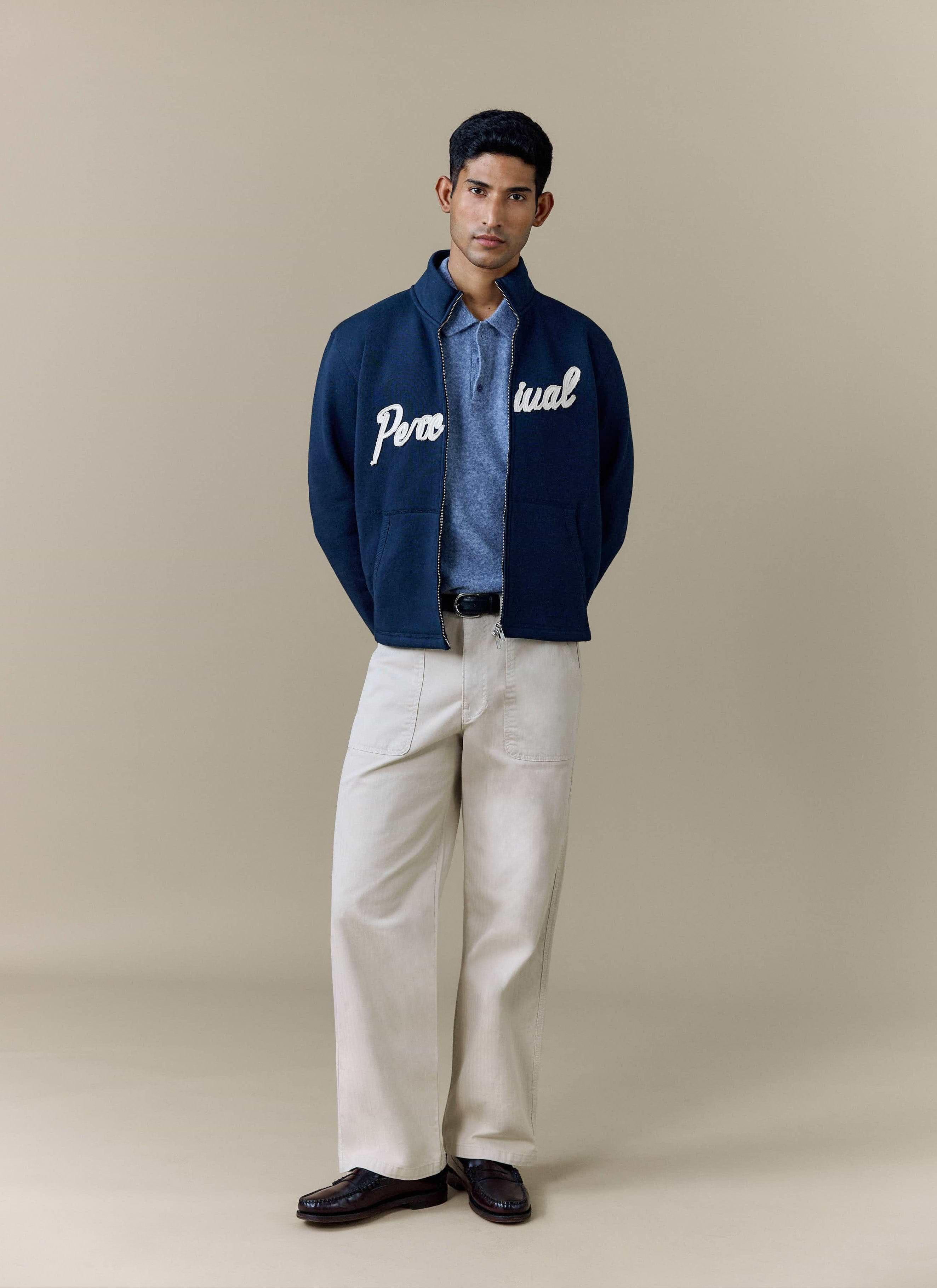 A model wears the navy Curtis Boxy Harrington from Percival, styled with a blue polo shirt and beige trousers.