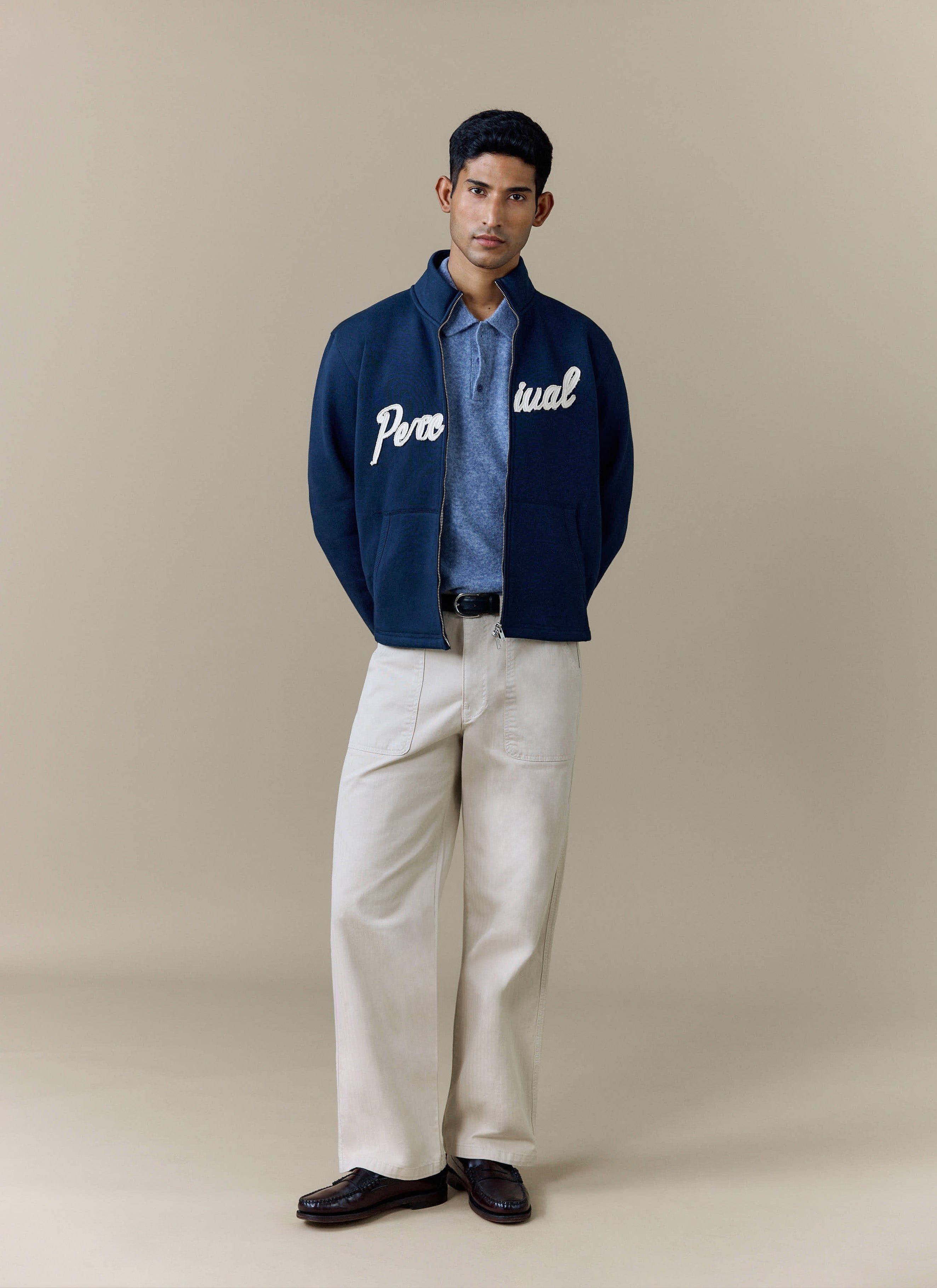 A model wears the navy Curtis Boxy Harrington from Percival, styled with a blue polo shirt and beige trousers.
