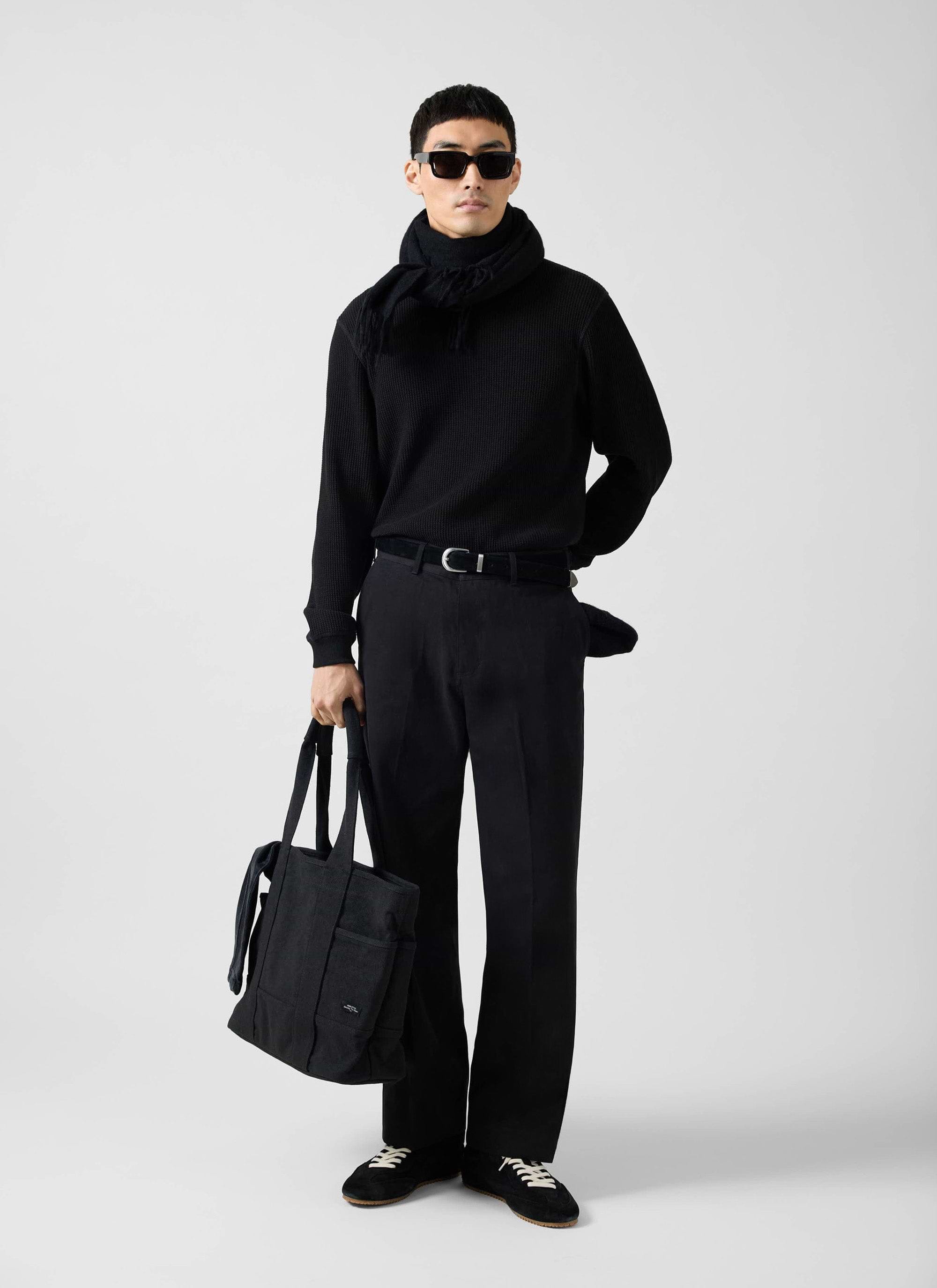 A model wears the black Waffle Long Sleeve T-Shirt from Percival, styled with black trousers, a scarf and sunglasses.