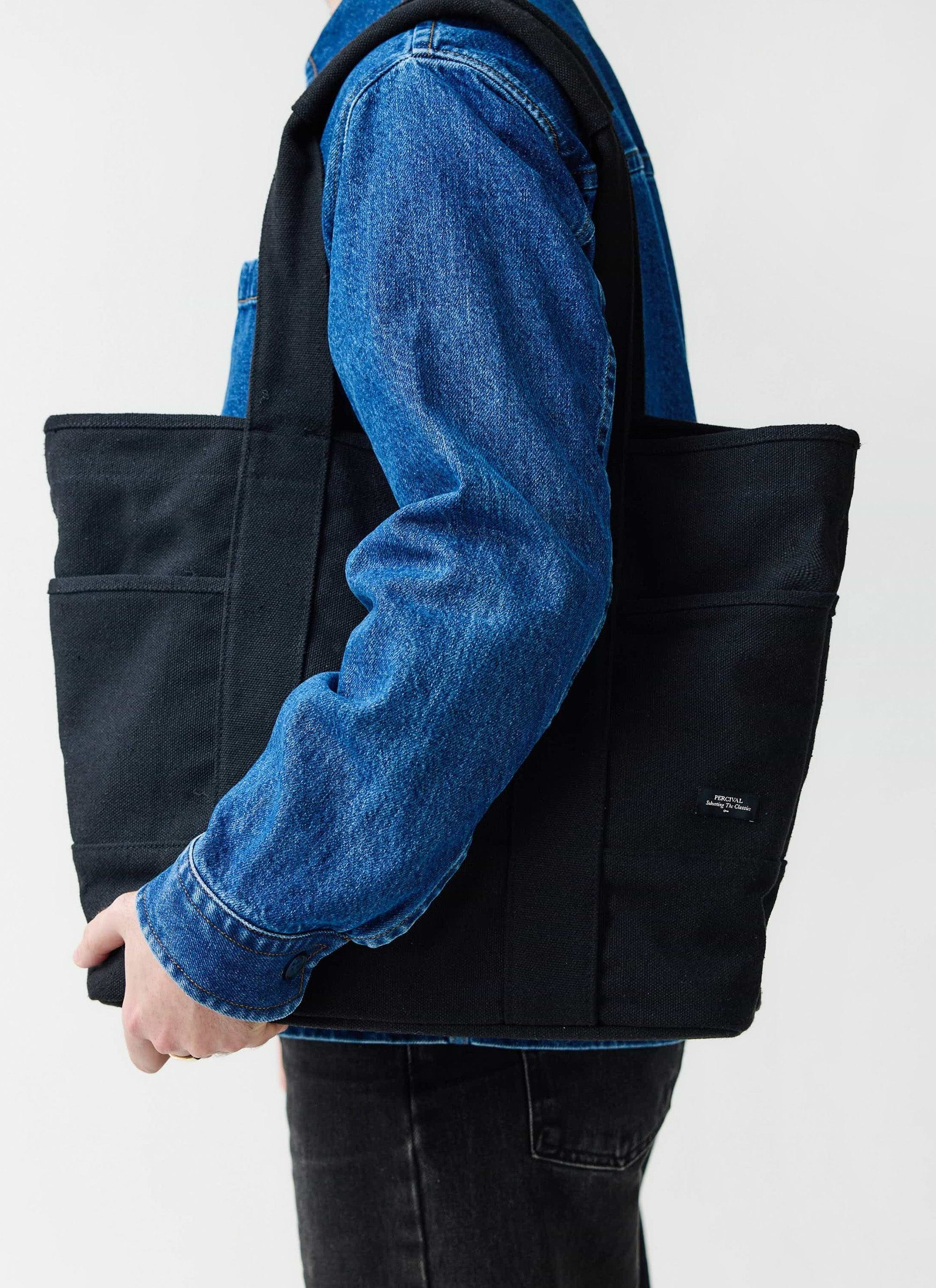 A model wearing a blue denim jacket carries the large black canvas tote bag from Percival.