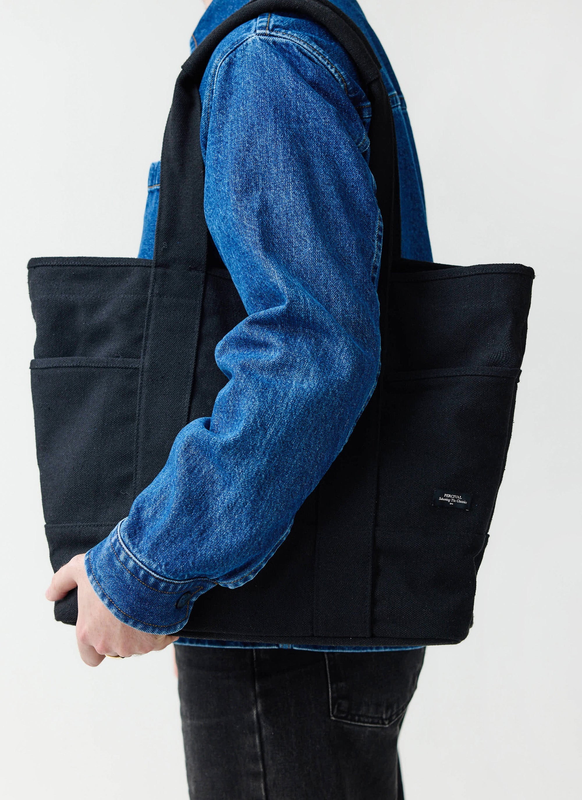 A model wearing a blue denim jacket carries the large black canvas tote bag from Percival.