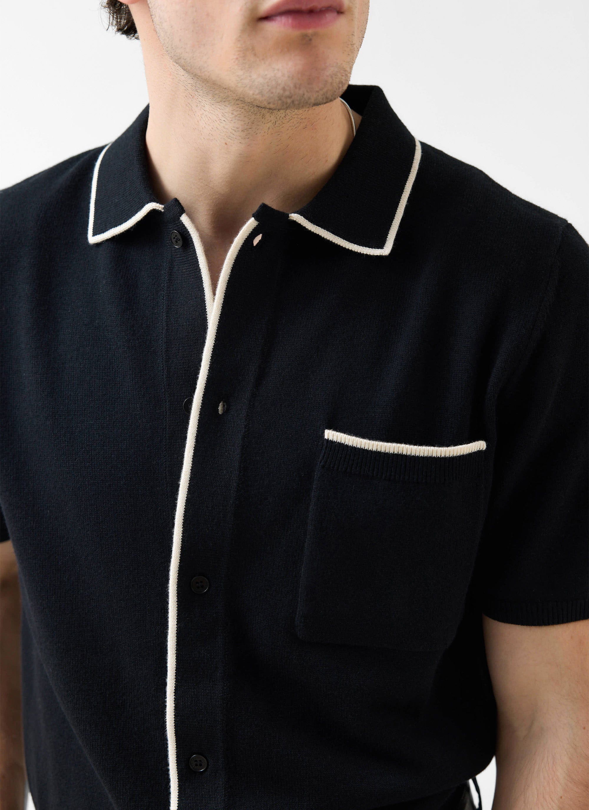 A close-up of a model wearing the Percival Contrast Trim Knitted Shirt in black with ecru detailing on the collar and pocket.