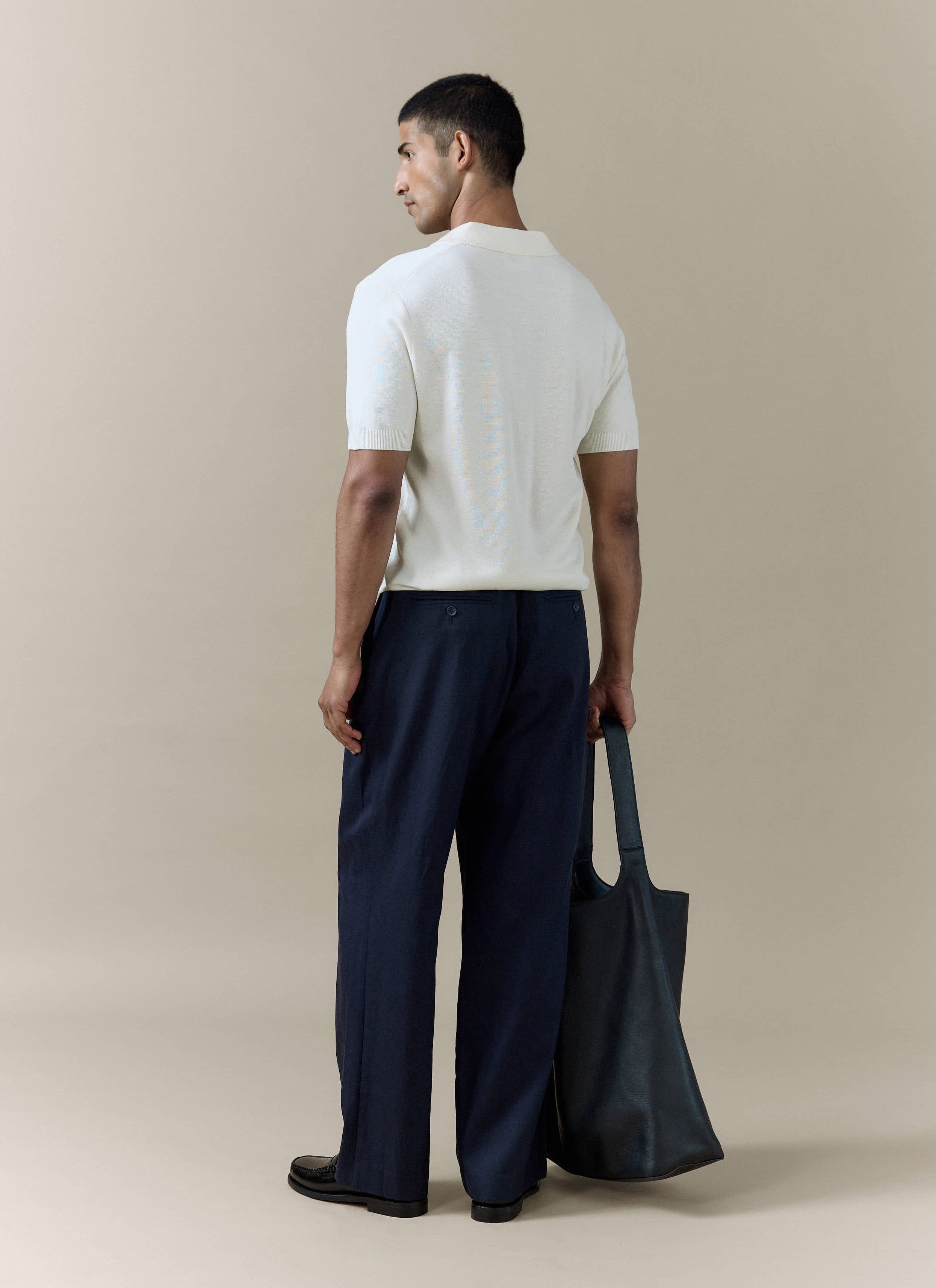 Rear view of the cream Laurence Knitted Polo by Percival in a linen blend, styled with navy trousers and a black tote.