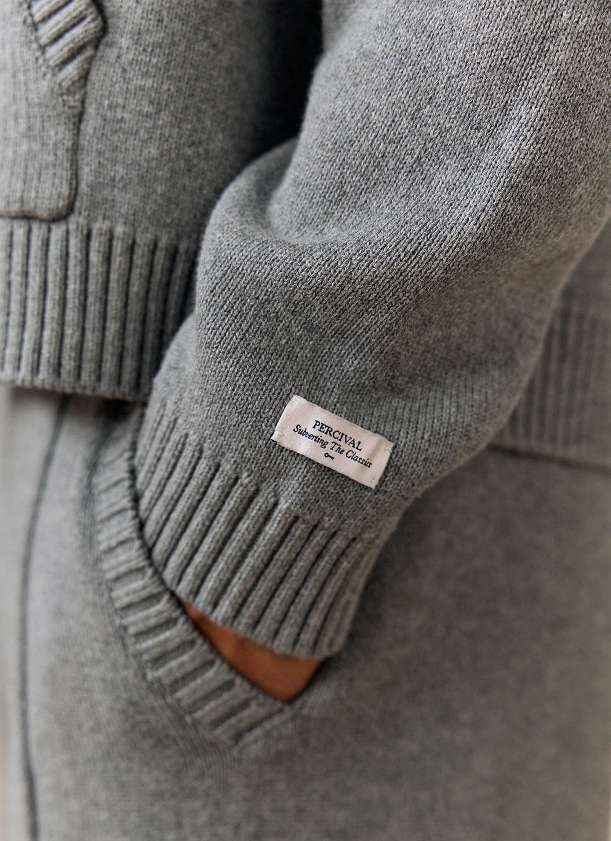 A close-up of the Percival woven brand label on the ribbed cuff of the grey cotton Chronicle Knitted Hoodie.