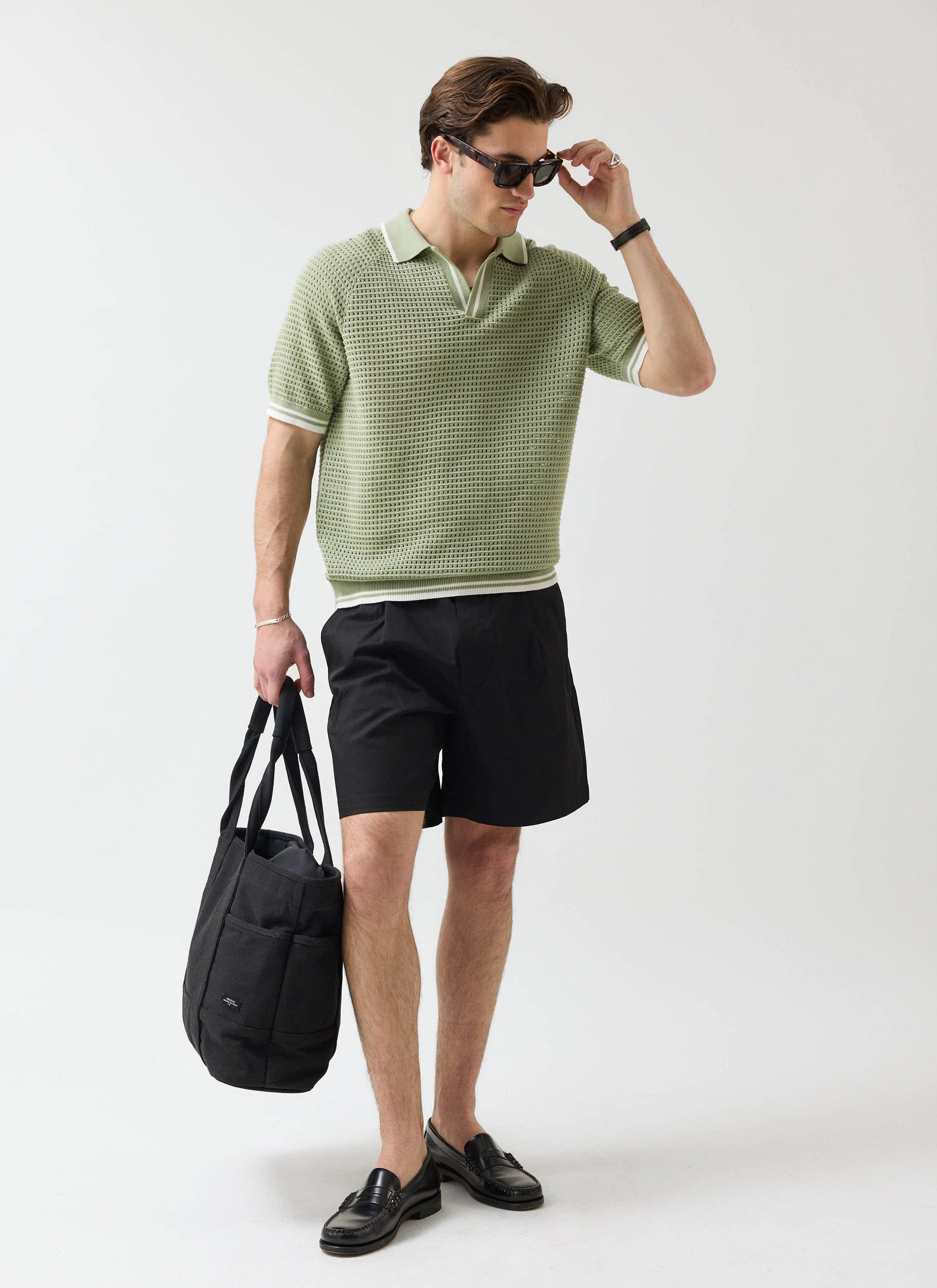 A model wearing the Percival sage knitted tennis polo, black shorts, and sunglasses, holding a large black tote bag.