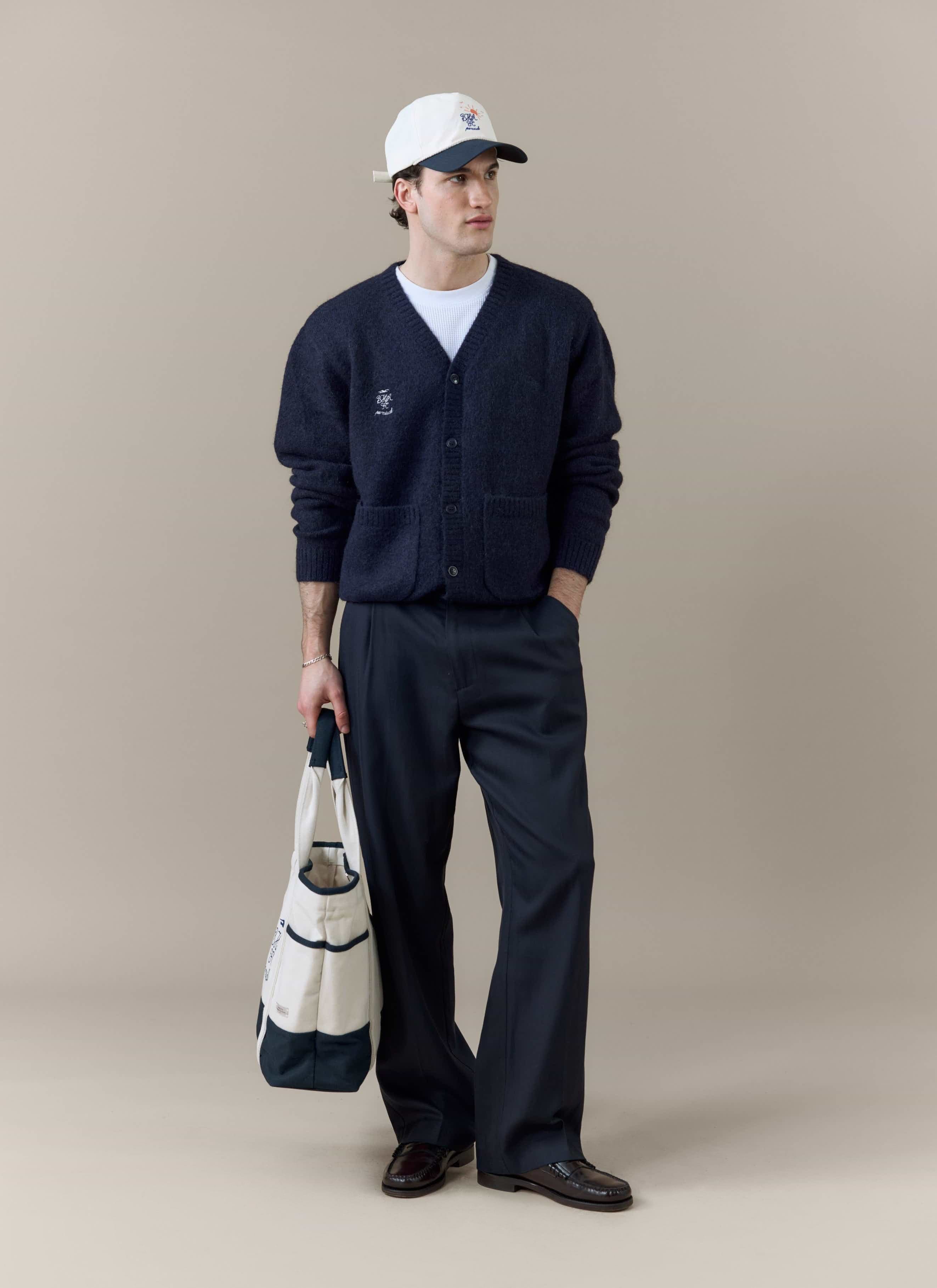 A model wears the navy Percival x Brighton & Hove Albion F.C. White Glove Cardigan, styled with trousers and a cap.