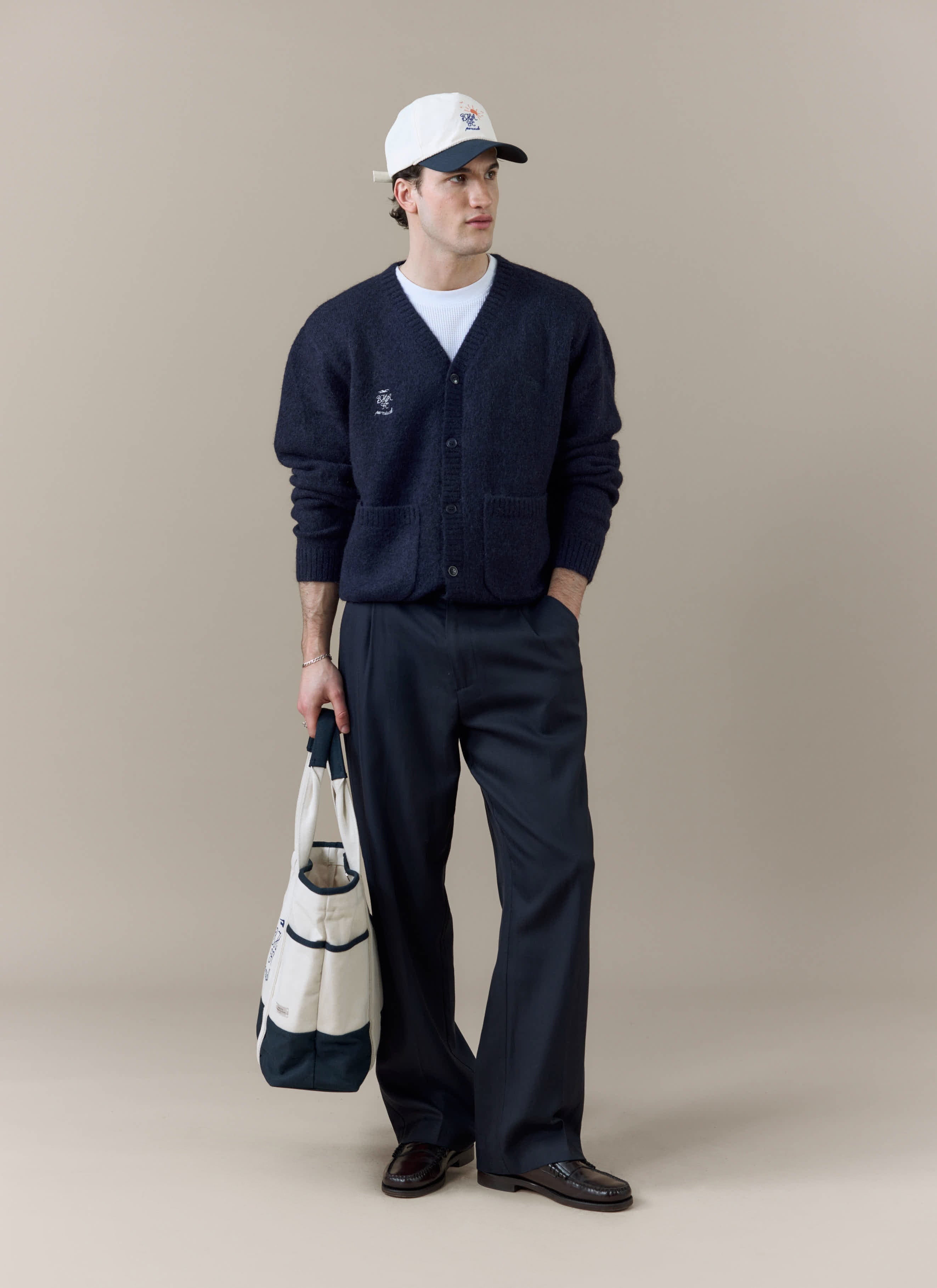 A model wears the navy Percival x Brighton & Hove Albion F.C. White Glove Cardigan, styled with trousers and a cap.