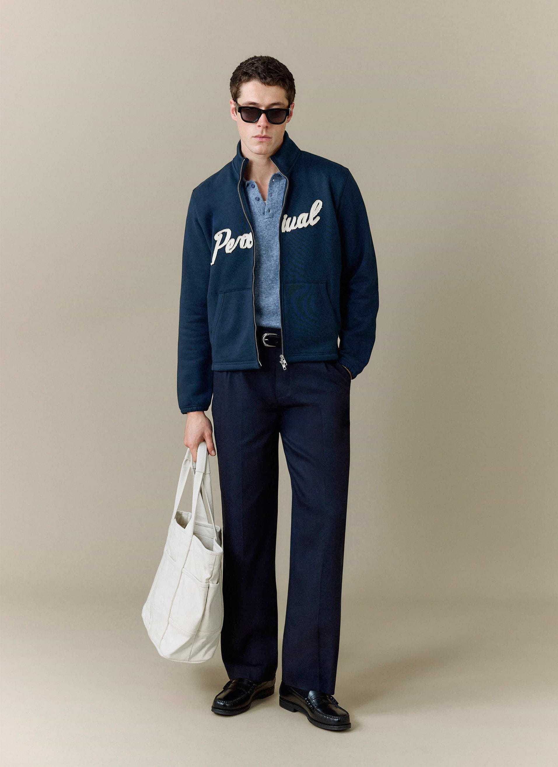 A model styles the Percival Collins Knitted Polo in blue under a navy logo jacket, with smart trousers and a white tote bag.