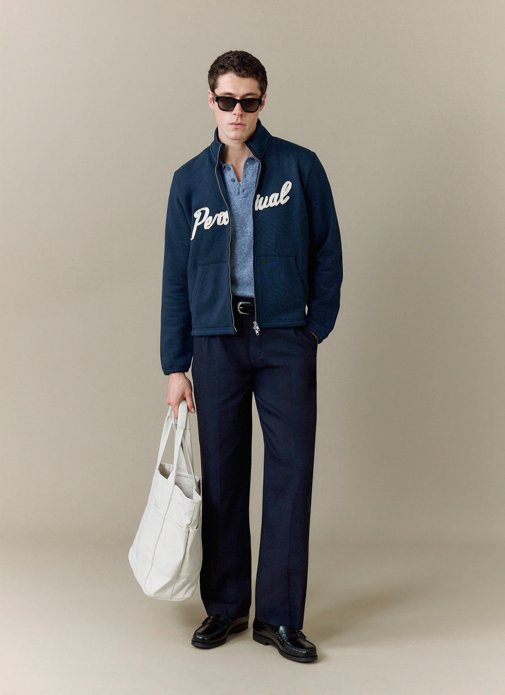A model styles the Percival Collins Knitted Polo in blue under a navy logo jacket, with smart trousers and a white tote bag.