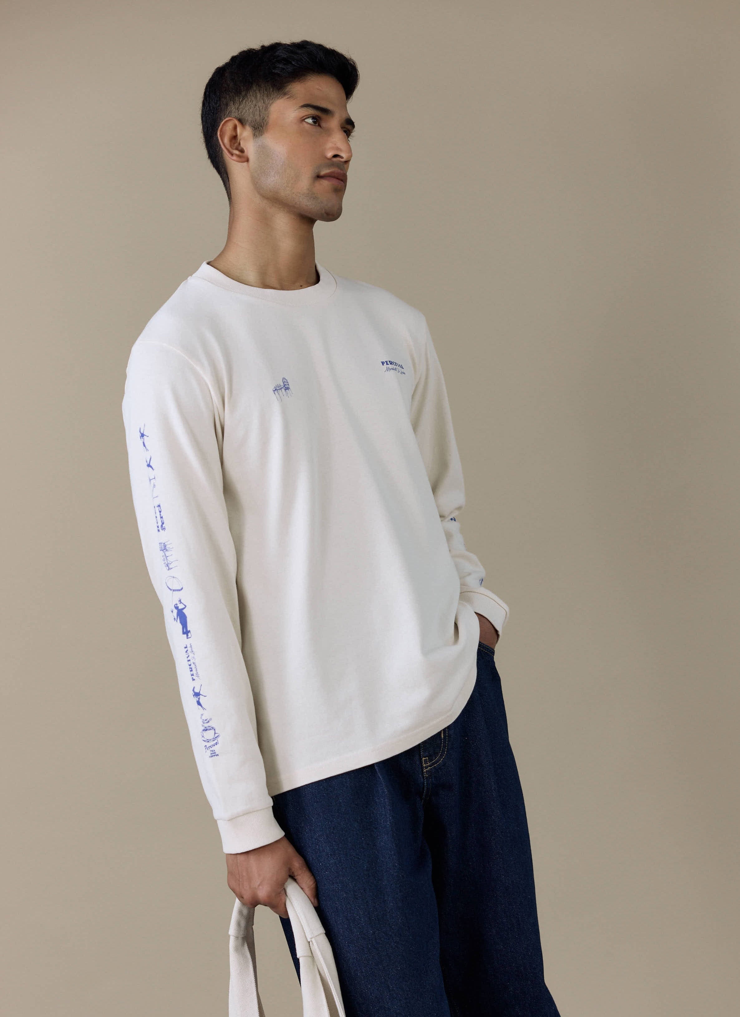 A model wearing the cream Harlow Fine Wines Long Sleeve T-Shirt by Percival with blue graphics on the sleeve and chest.