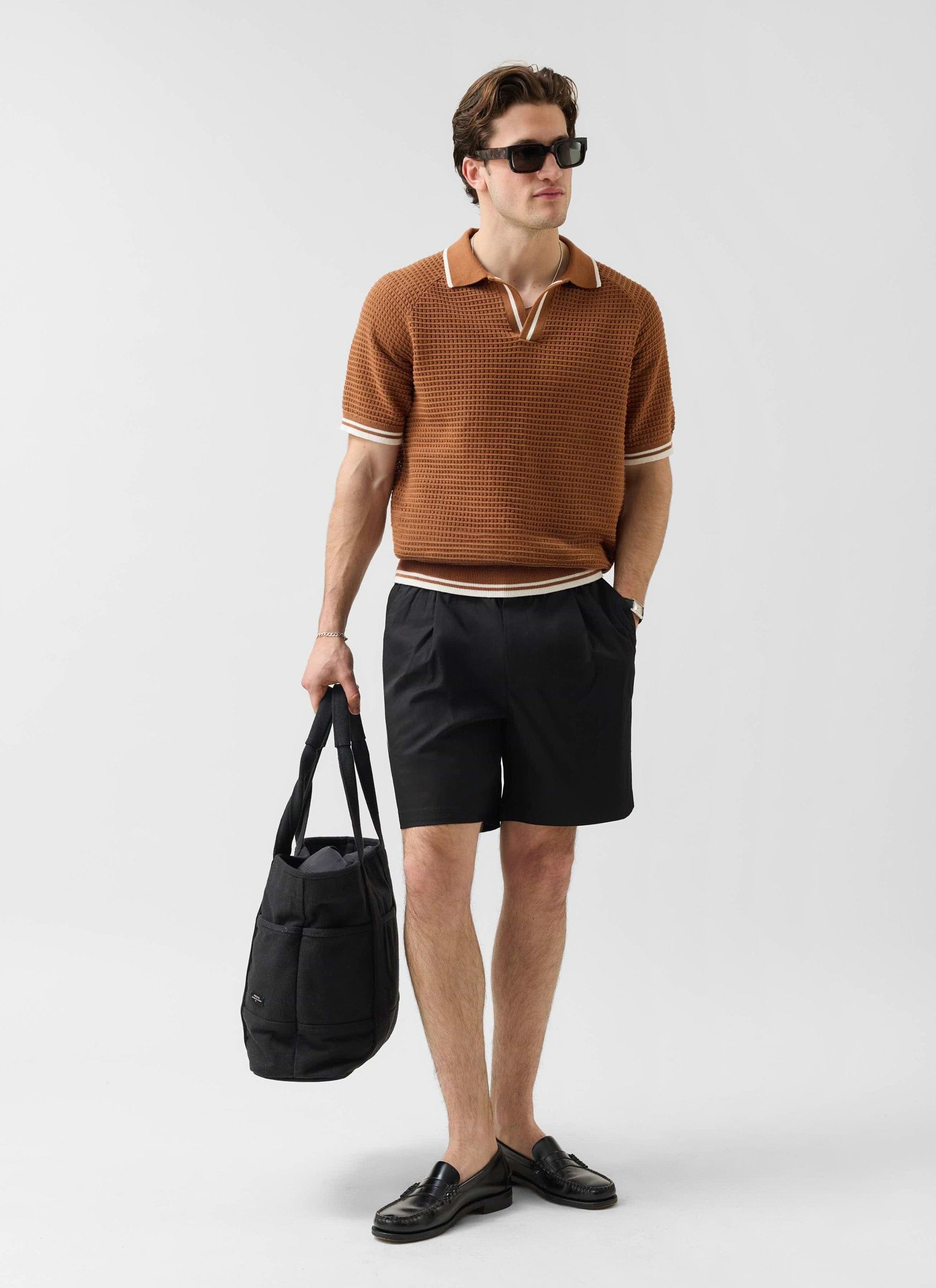 A man models the brown Percival Tennis Boxy Knitted Polo, styled with black shorts, loafers, and a large black tote bag.