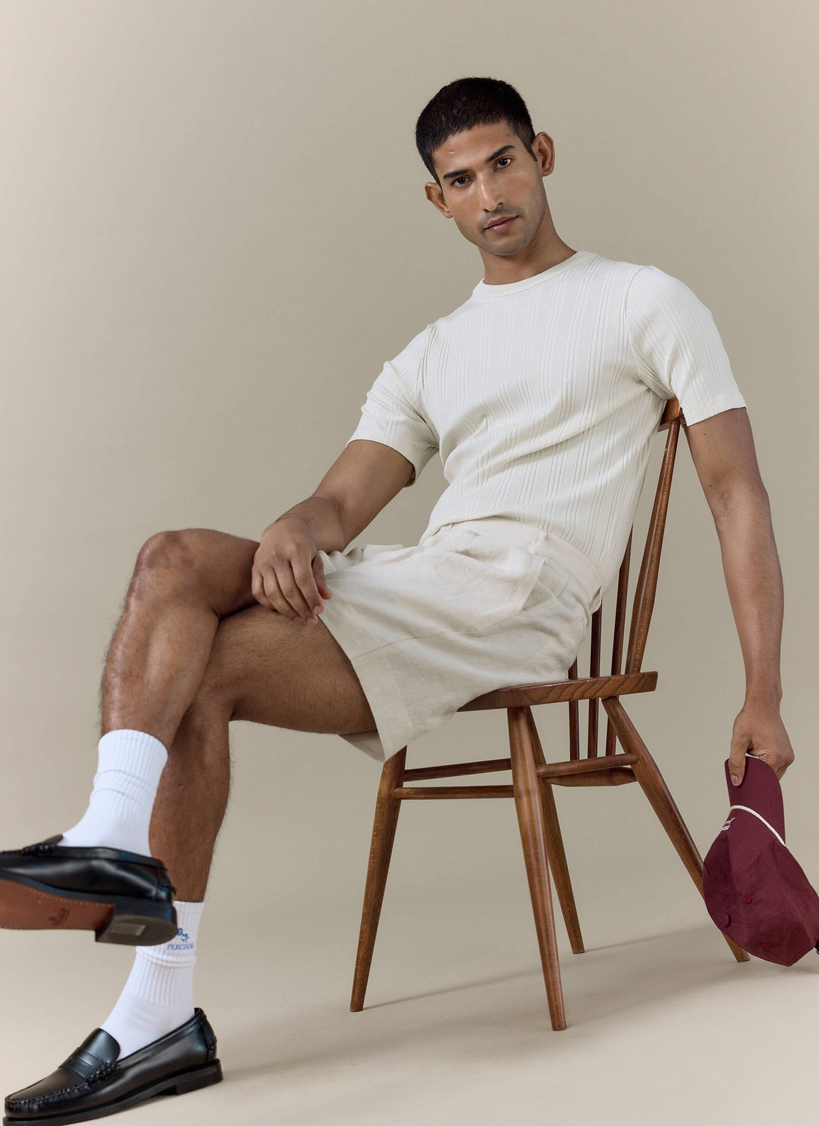 Percival pleated linen shorts in cream worn with a matching ribbed t-shirt by a model sitting on a wooden chair.