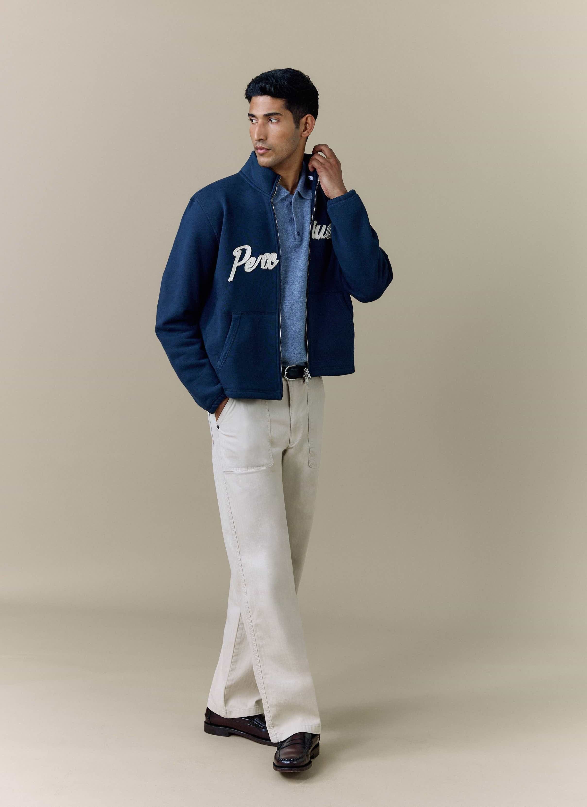 A model wears the Percival Curtis Boxy Harrington in navy, layered over a blue polo shirt and styled with cream trousers.