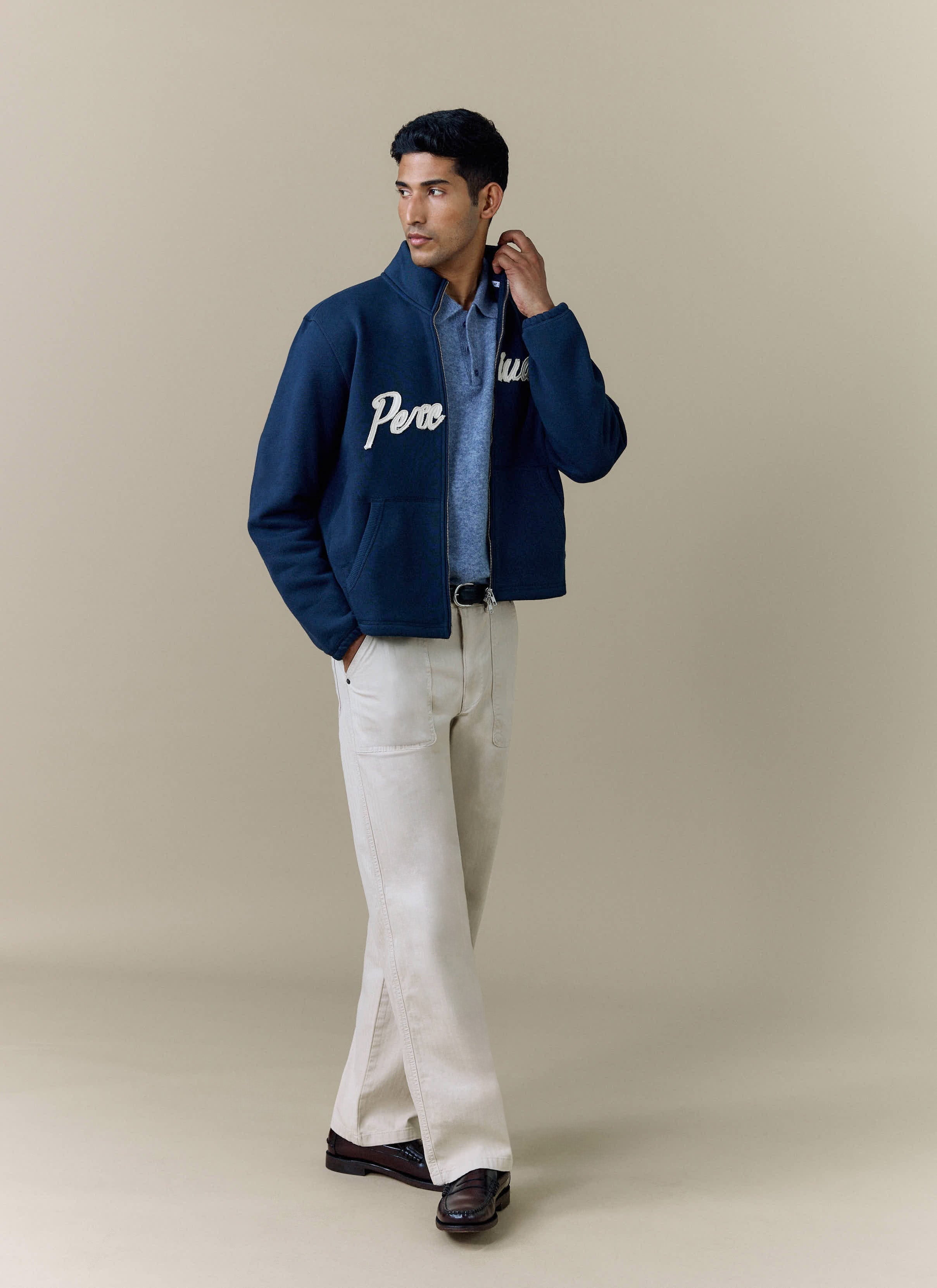 A model wears the Percival Curtis Boxy Harrington in navy, layered over a blue polo shirt and styled with cream trousers.