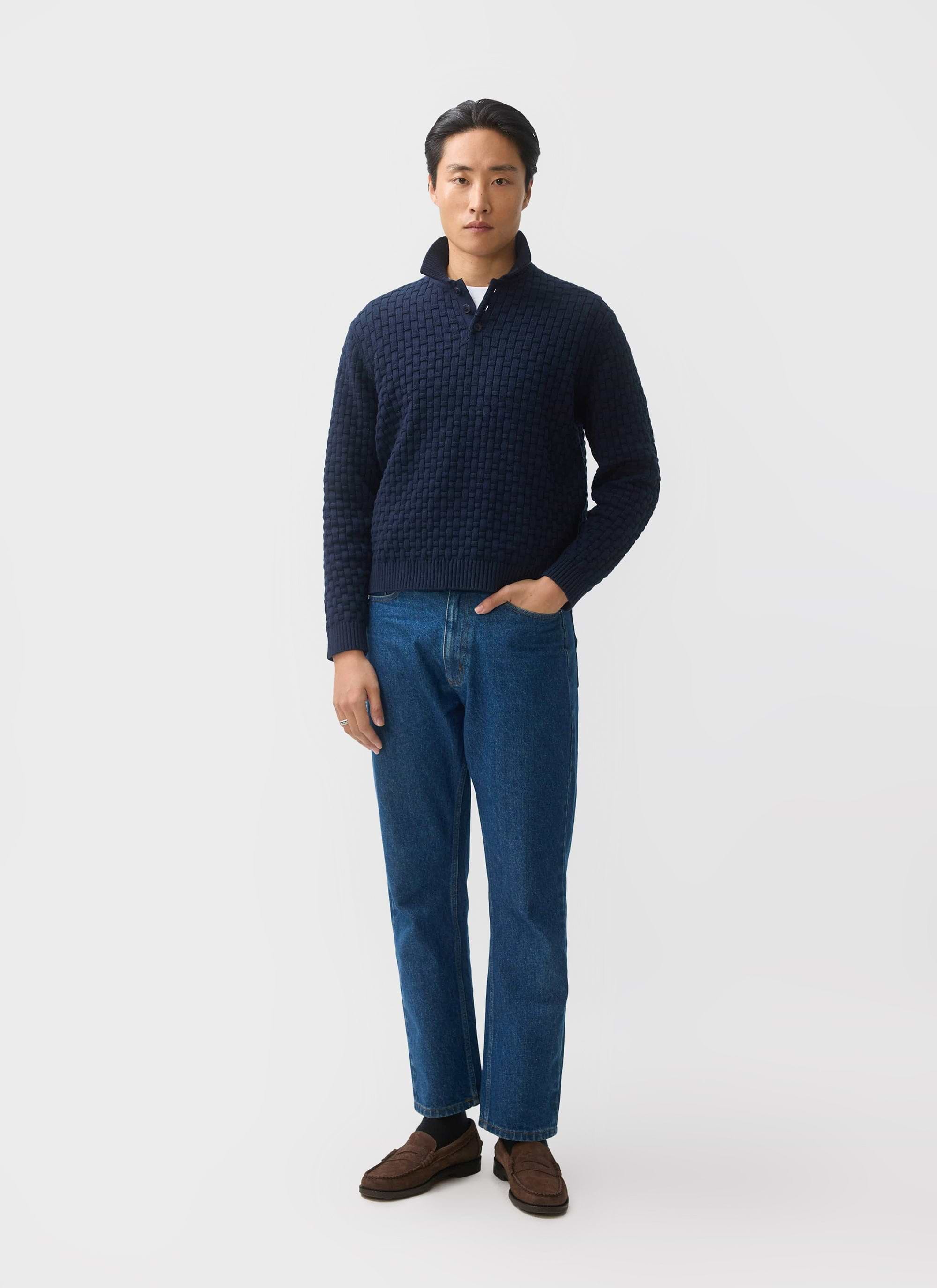 A man models the Percival Archer Basketweave Knitted Shirt in navy, styled with blue jeans and brown loafers.
