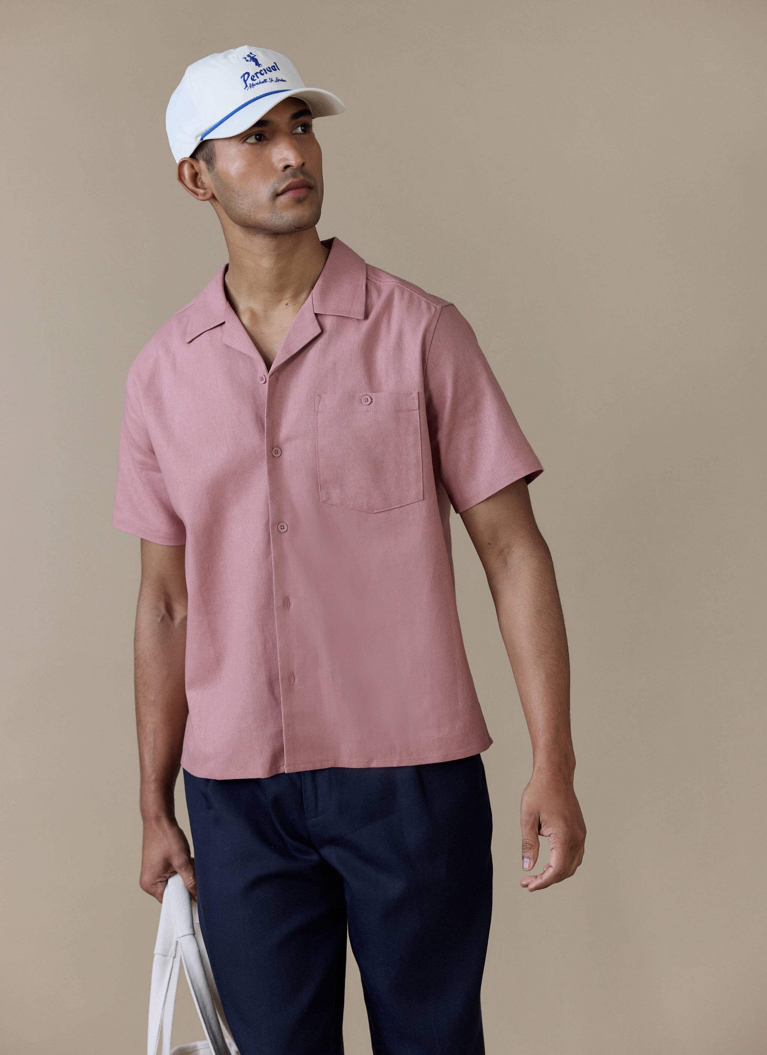 A man wearing the Percival Short Sleeve Cuban Shirt in dusty pink linen blend with navy trousers and a white Percival cap.