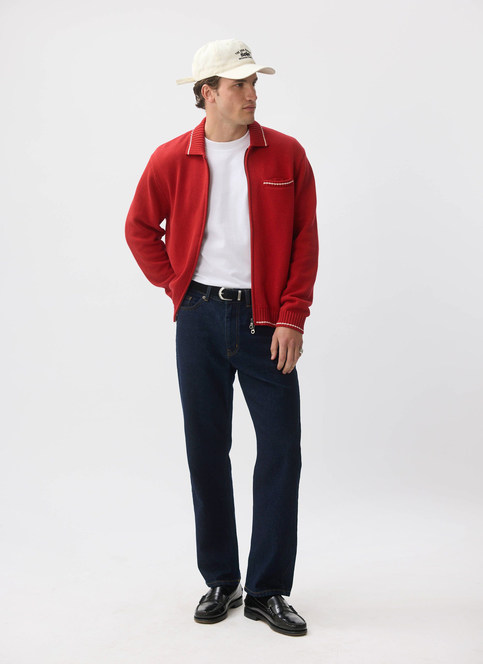 A man models the Percival Gardner Dense Knitted Zip Cardigan in red, styled over a white tee and dark denim jeans.