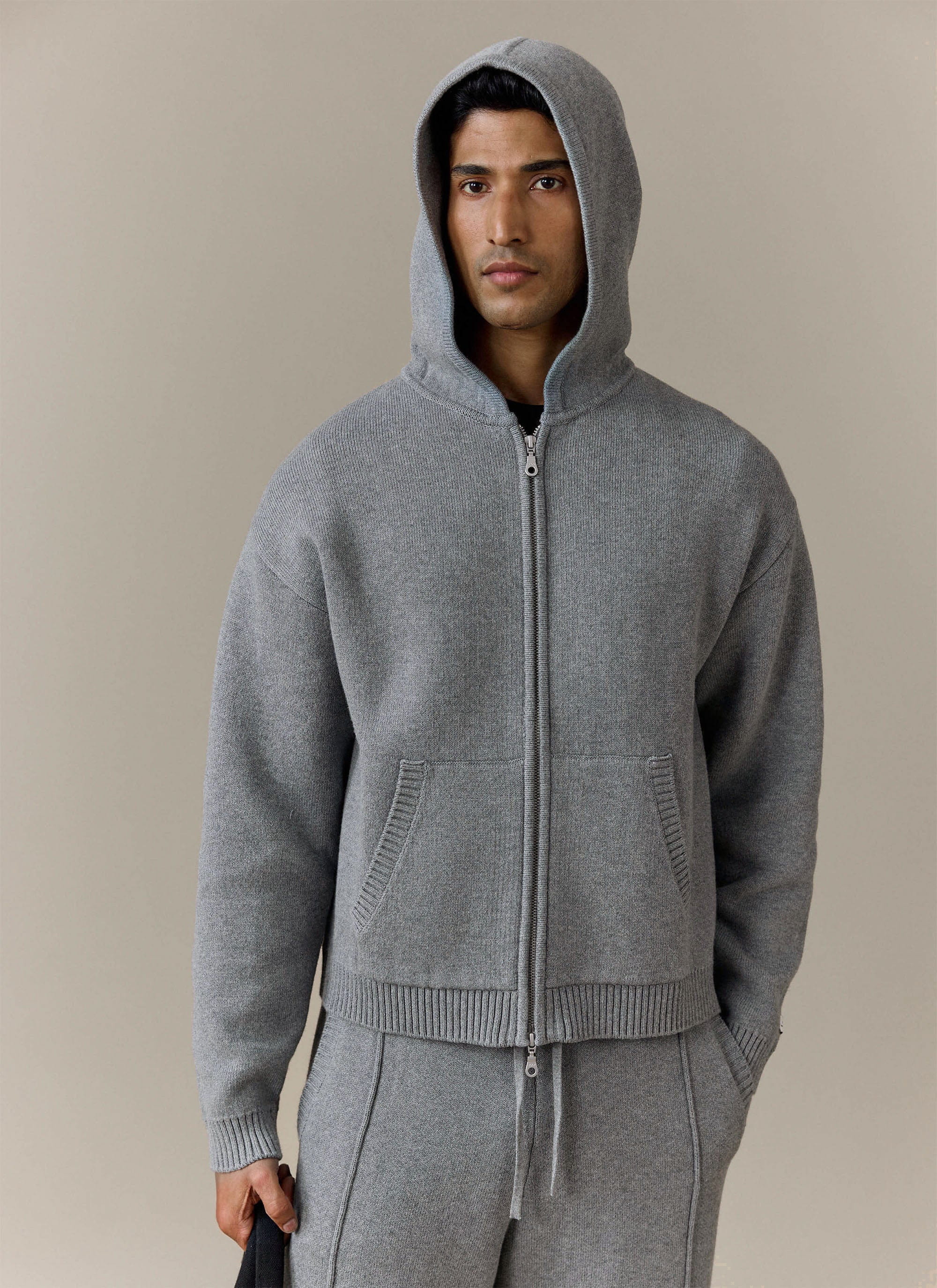 A model wears the grey Chronicle Knitted Hoodie from Percival with the hood up, looking directly at the camera.