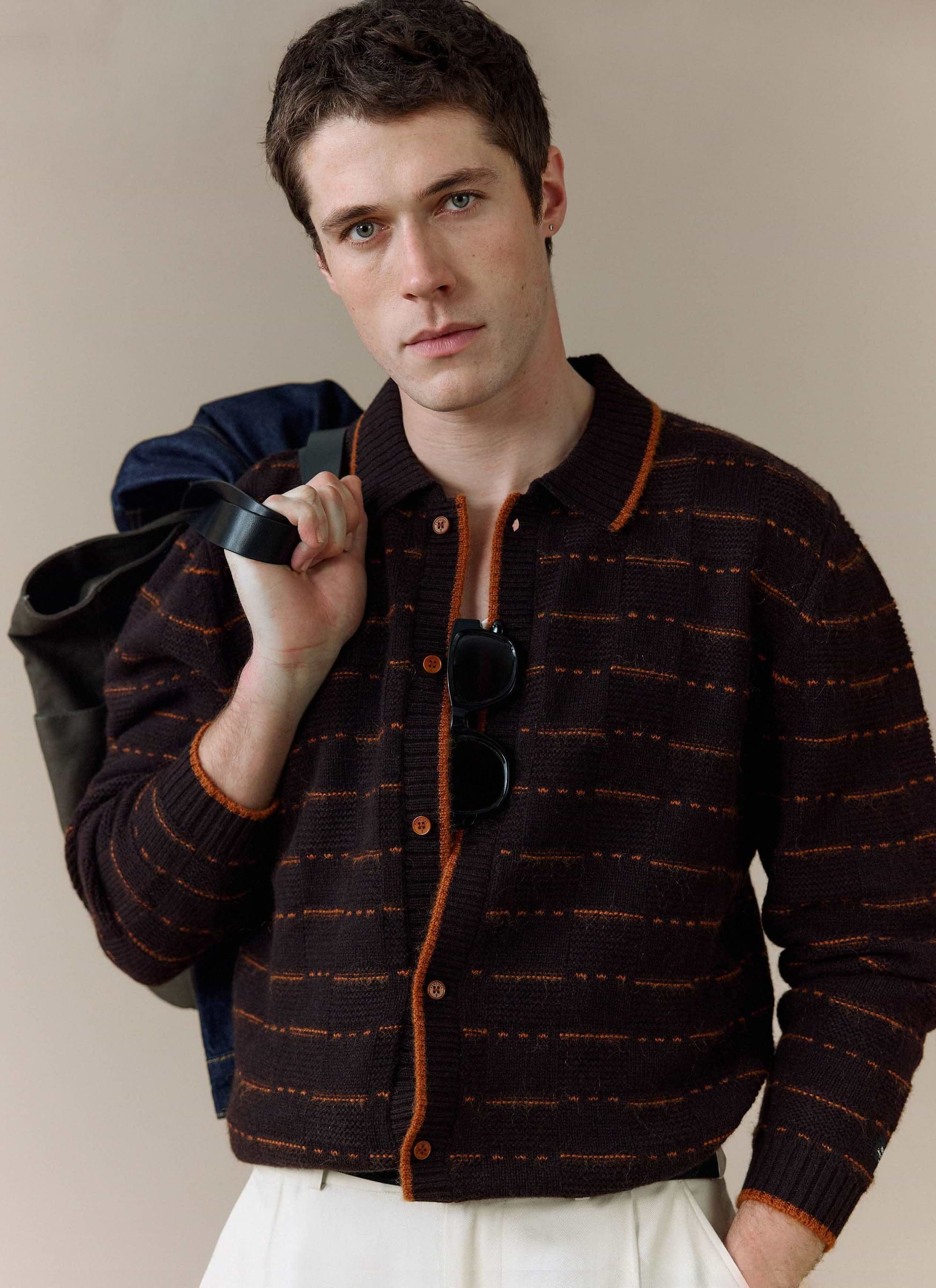 A man models the brown Addison Long Sleeve Knitted Shirt from Percival, styled with white trousers and carrying a bag.