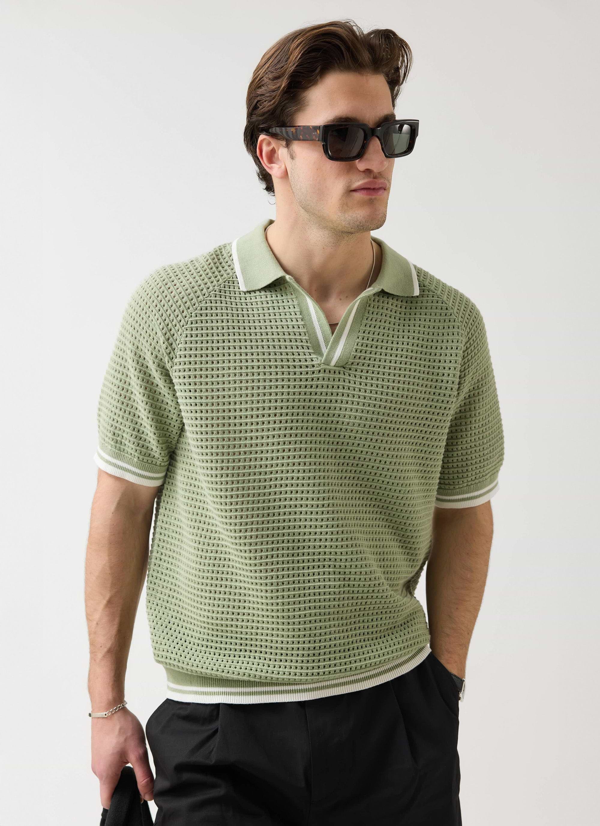 A man models the sage green Percival Tennis Boxy Knitted Polo with sunglasses against a white studio background.