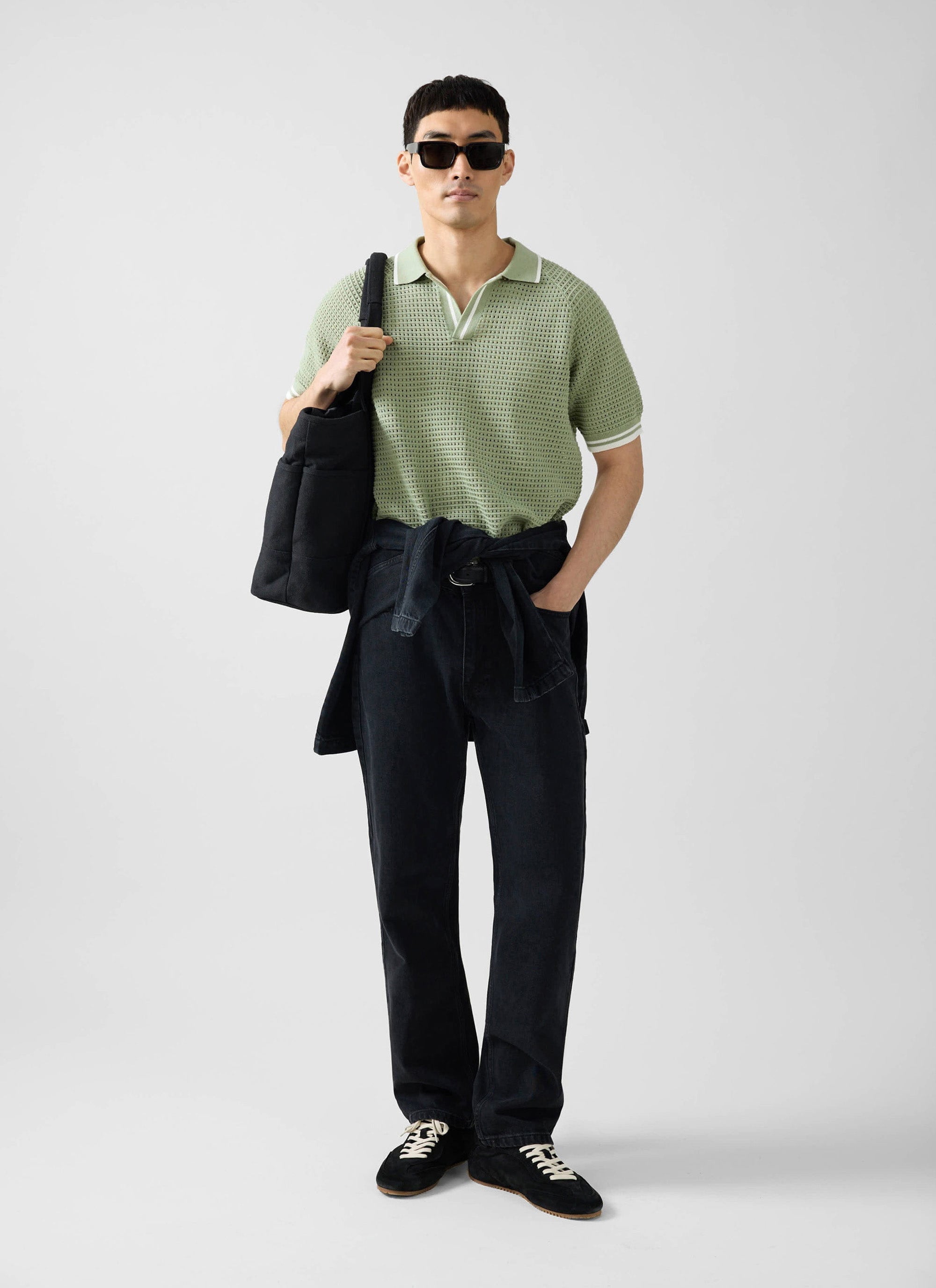 A man wearing the Percival sage green Tennis Boxy Knitted Polo, black jeans, sunglasses, and carrying a black tote bag.