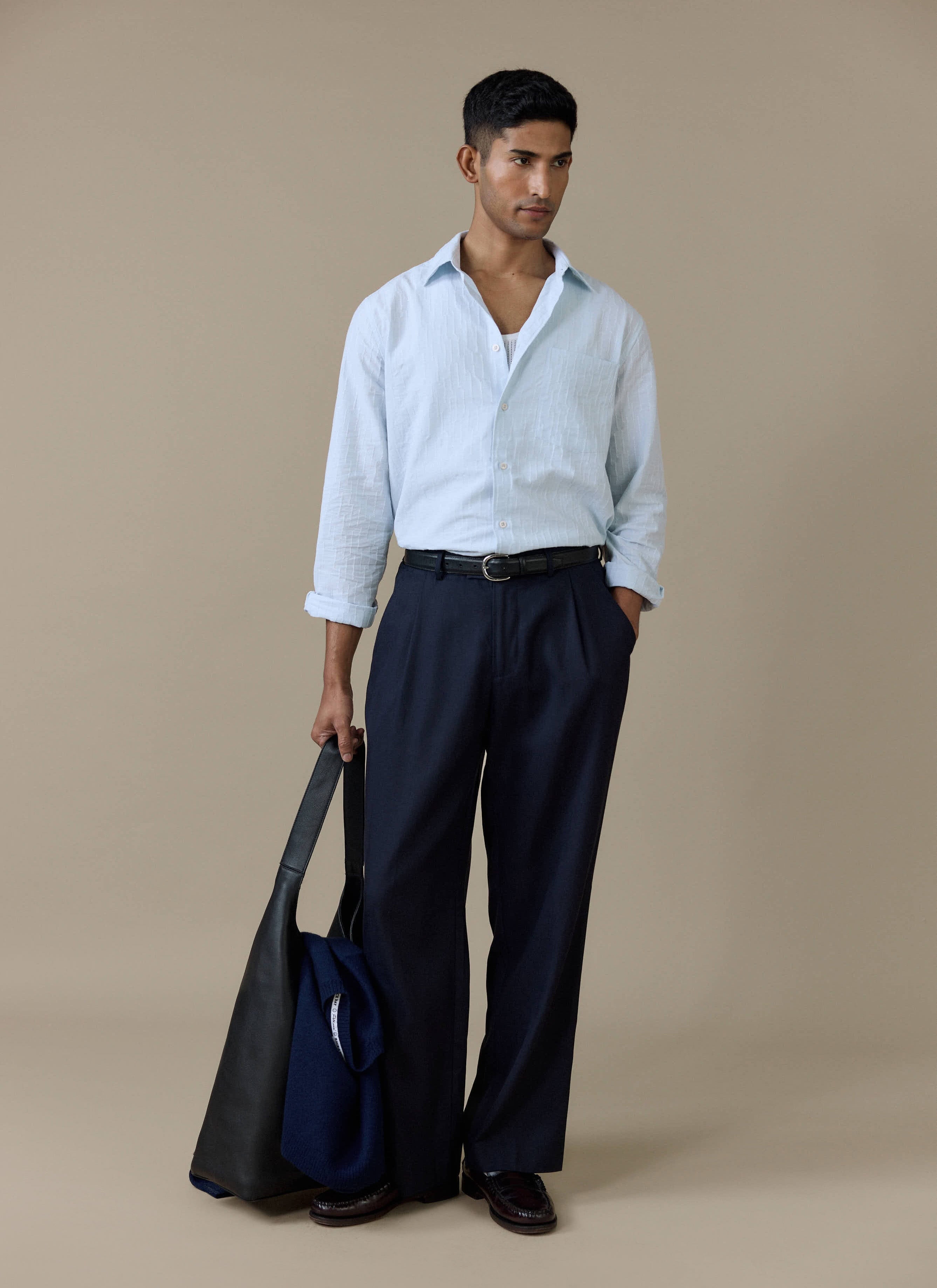 A model wearing the Percival Paros Pearce Shirt in light blue seersucker, styled with dark trousers and a black bag.
