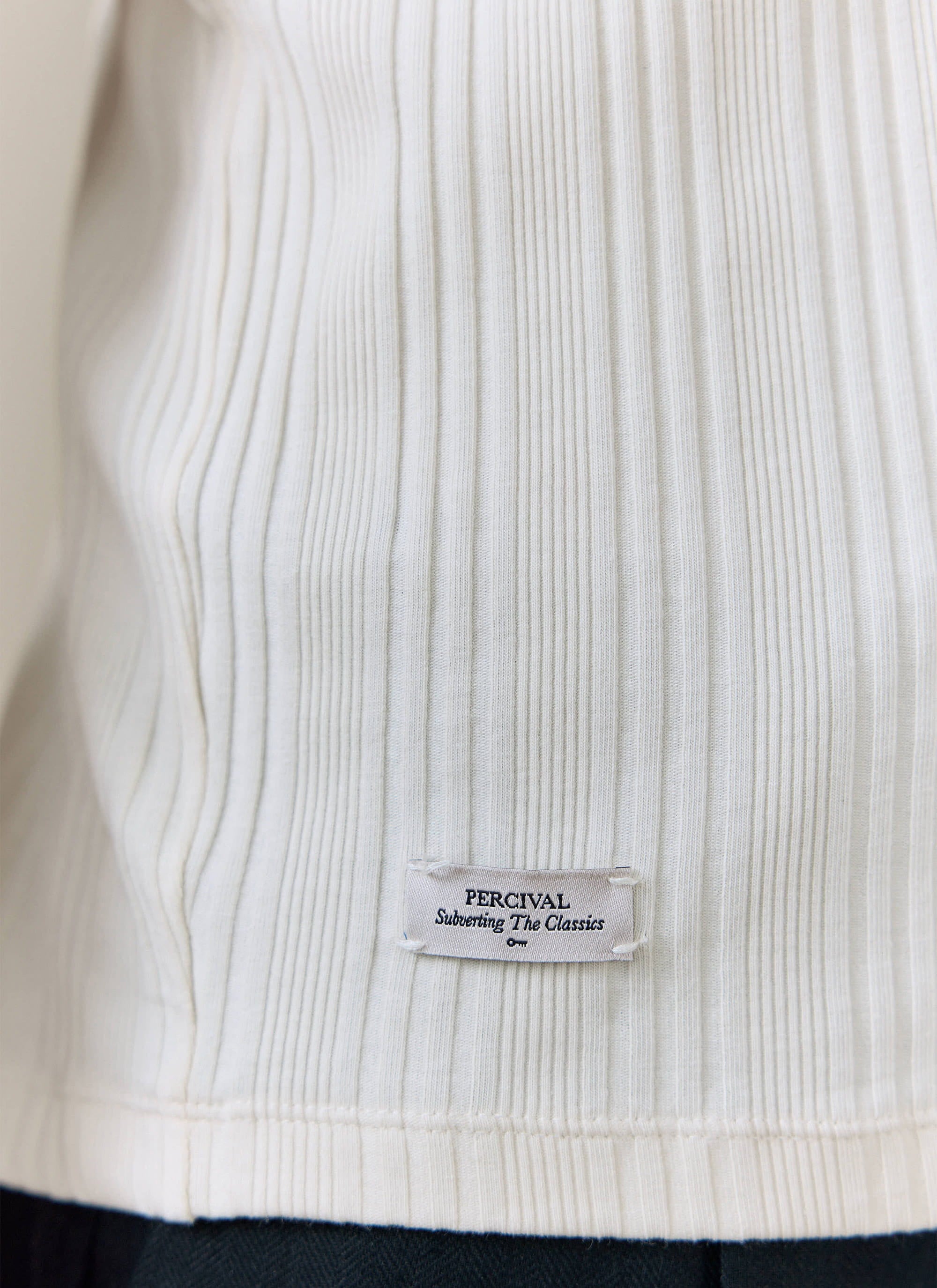 Close-up of the cream ribbed cotton Leon T-shirt by Percival, highlighting the branded 'Subverting The Classics' hem tag.