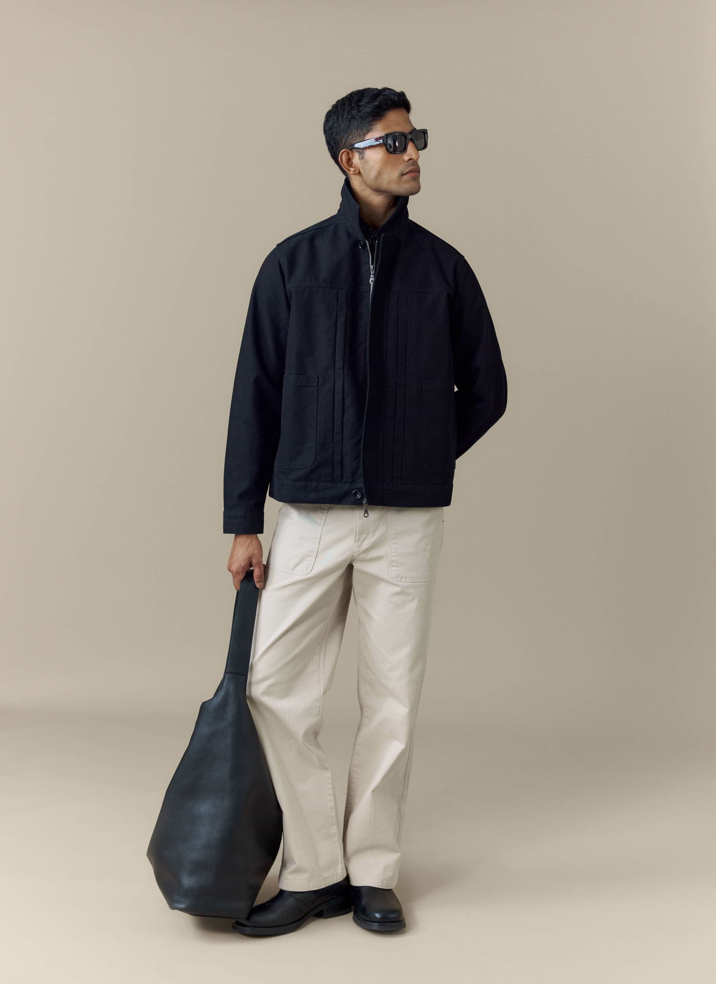 A model wears the black cotton Ernst Jacket from Percival, styled with cream trousers, sunglasses and a black bag.