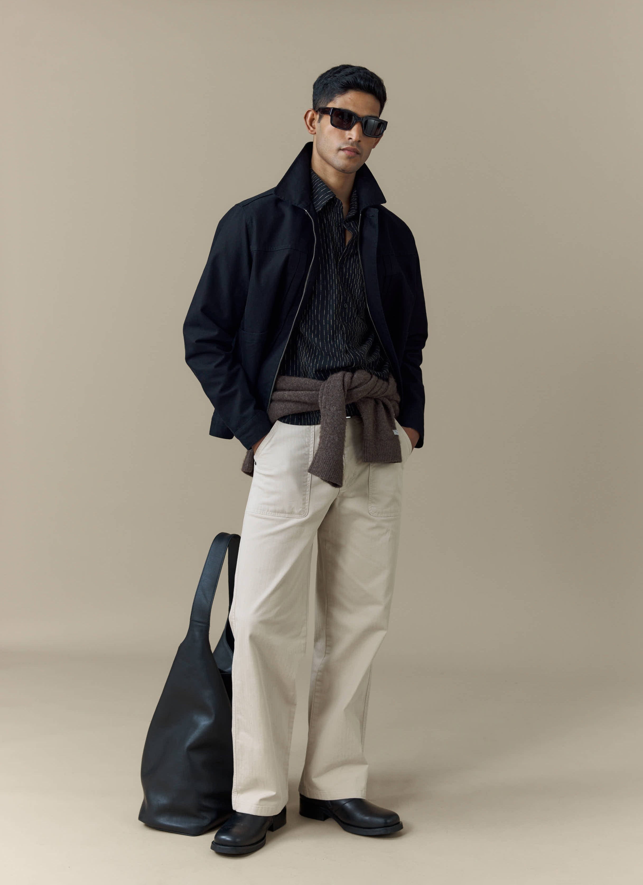 A model wears the black cotton Ernst Jacket from Percival, styled with a striped shirt and cream trousers.