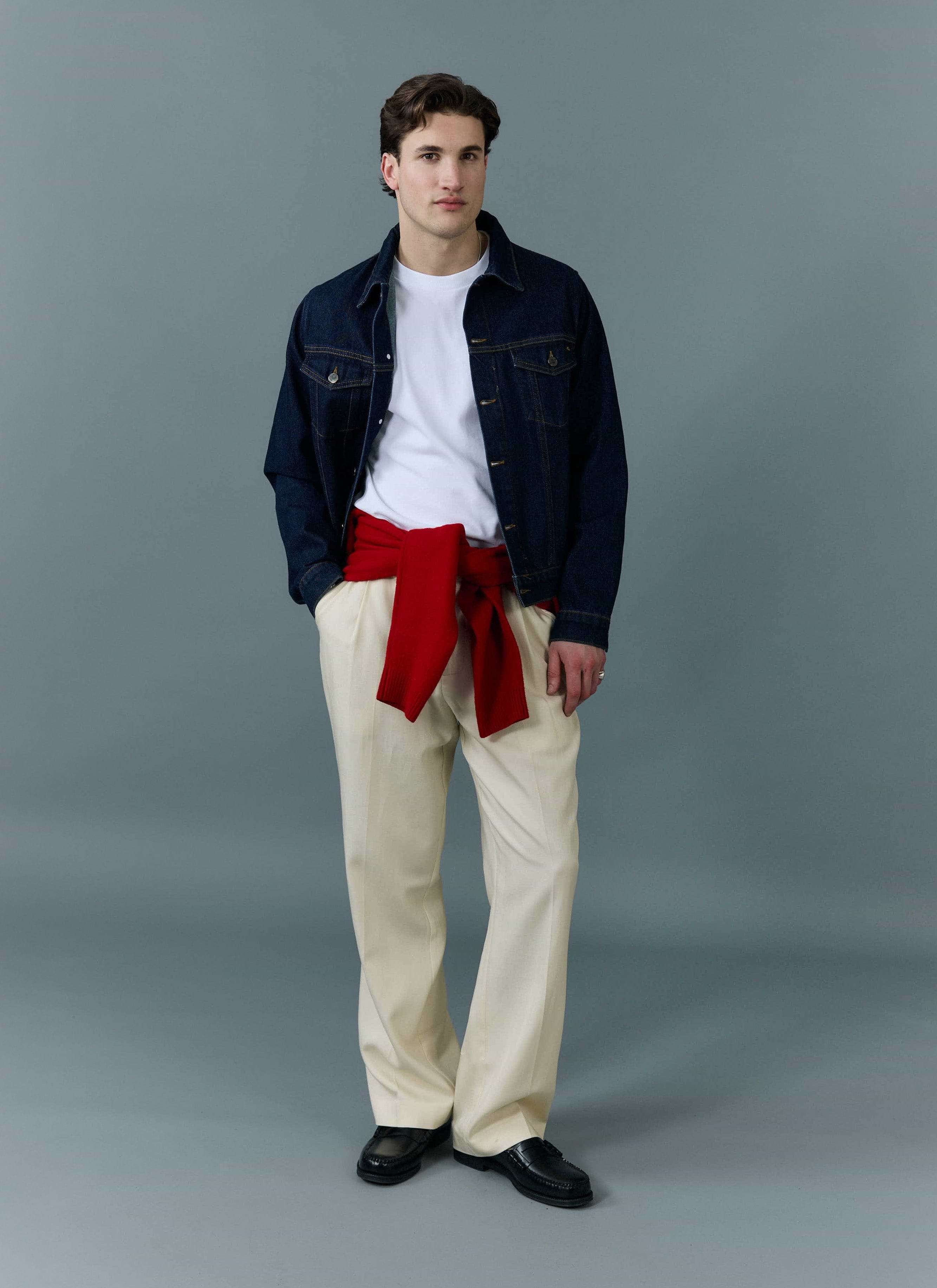A man models the Percival dark indigo Western Trucker Jacket, styled with a white t-shirt, cream trousers and a red jumper.