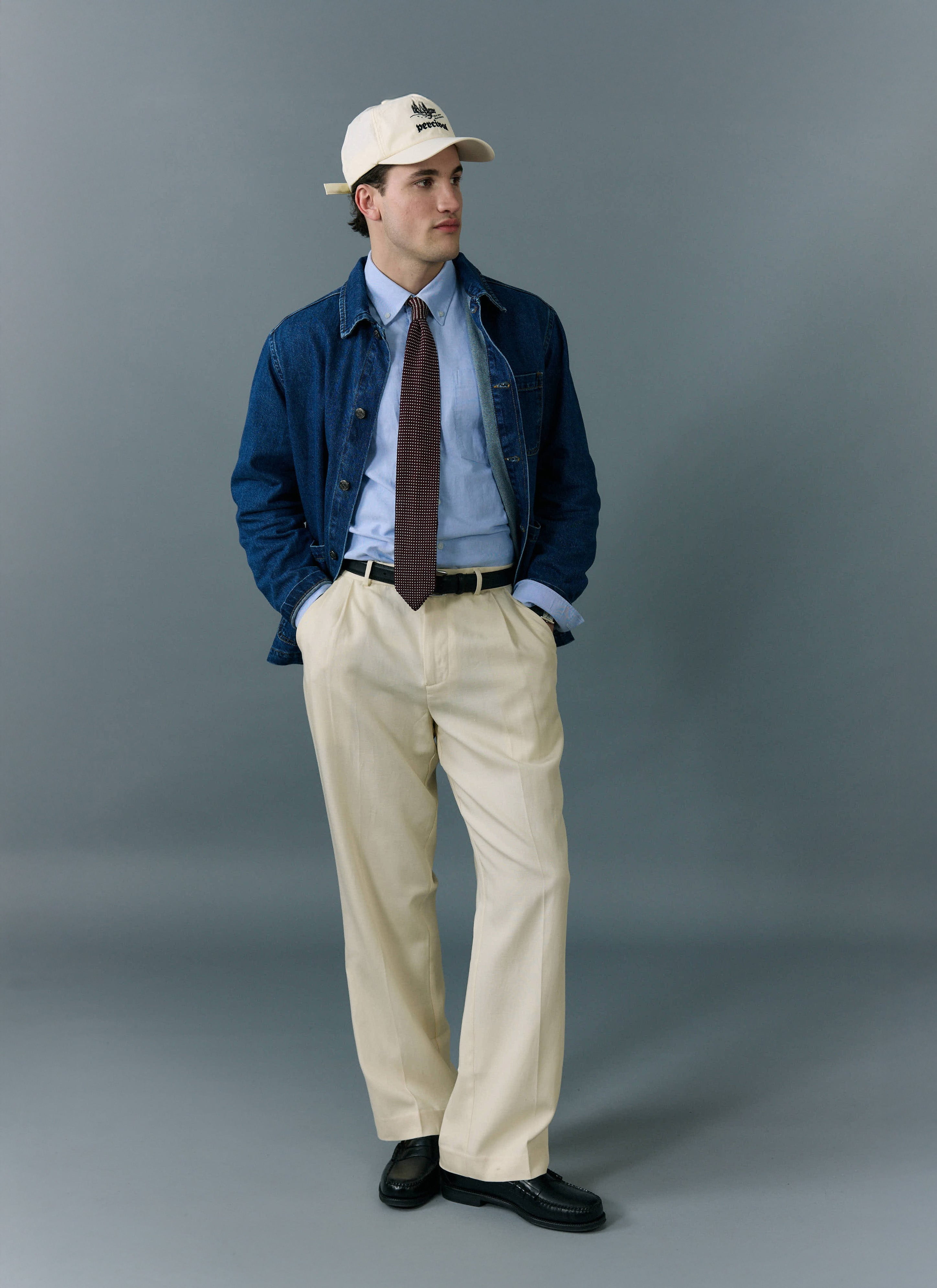 A model styles the Percival Gove Chore Jacket in mid wash blue with a shirt, tie, cream trousers and baseball cap.