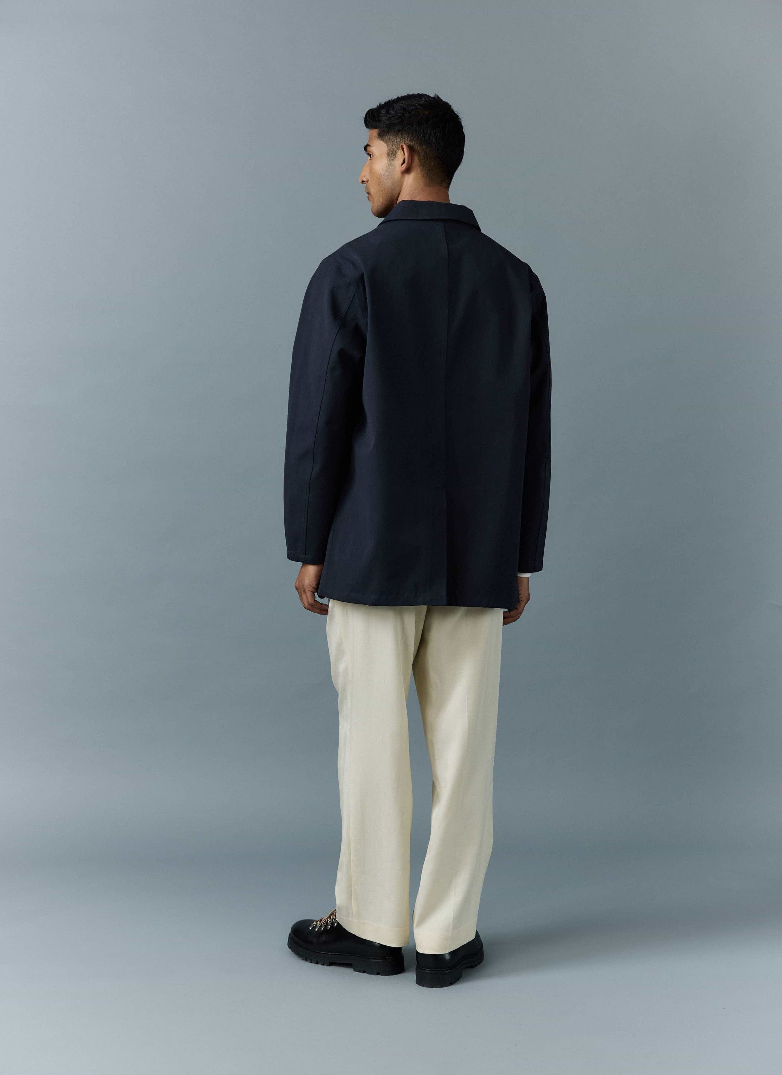 A model, seen from the back, wears the navy waterproof Sherlock Trench Coat from Percival with cream trousers.