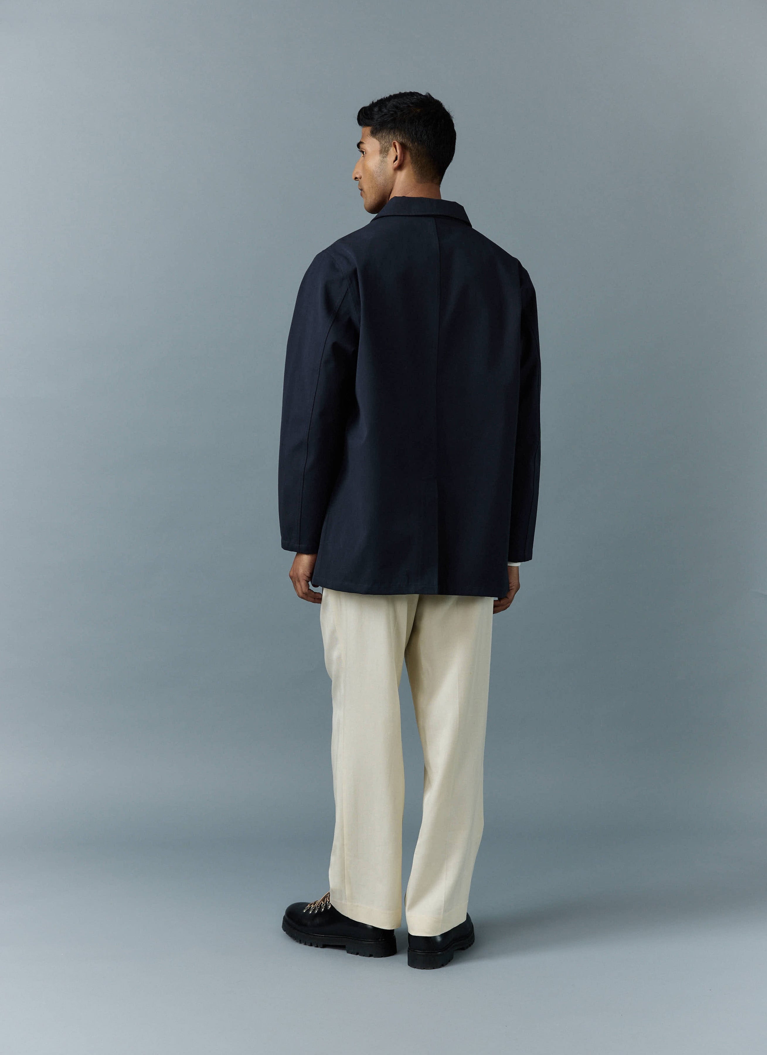 A model, seen from the back, wears the navy waterproof Sherlock Trench Coat from Percival with cream trousers.