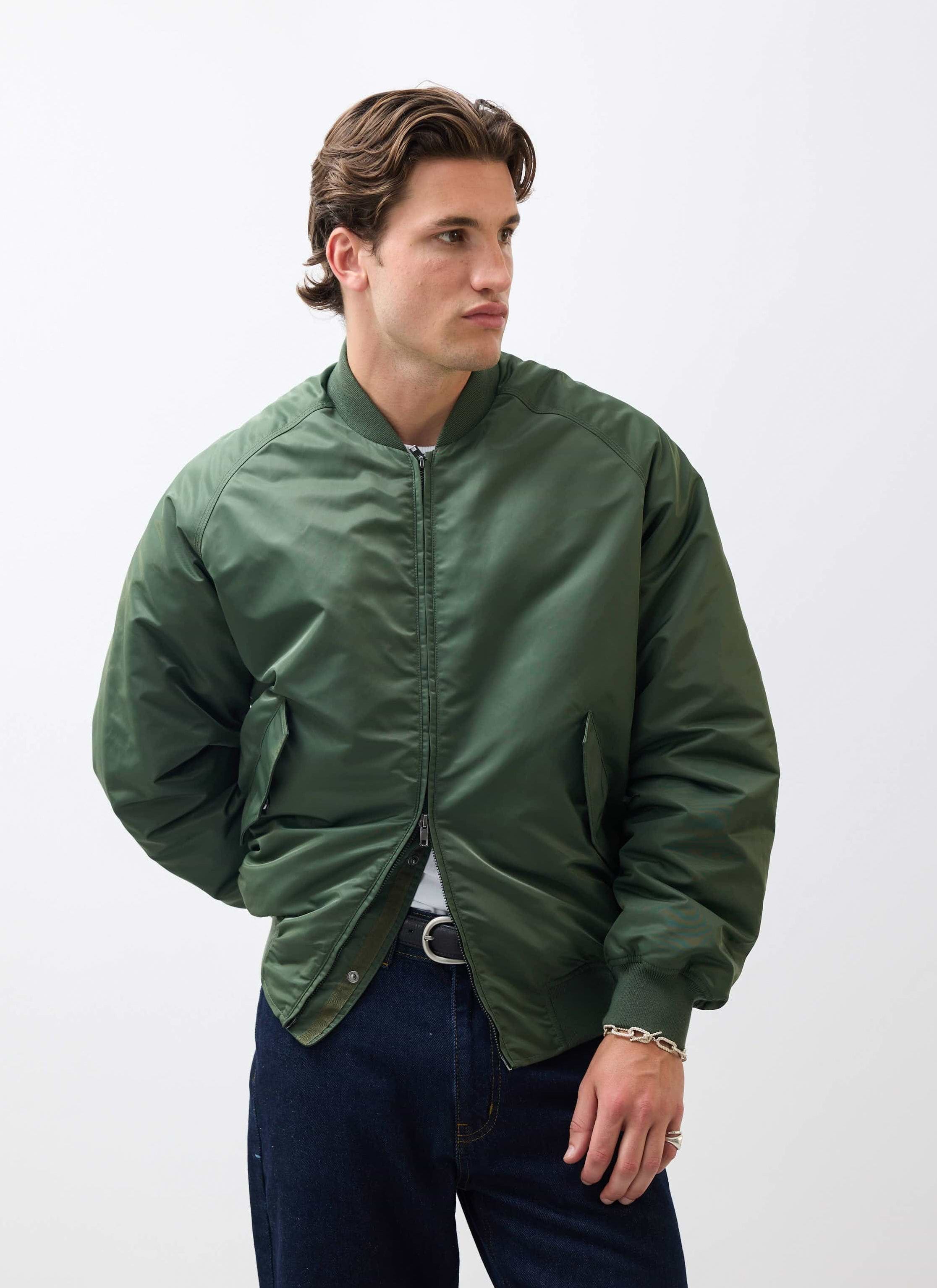 A model wears the Percival Bomber Jacket in forest green nylon against a white studio background, looking to the side.