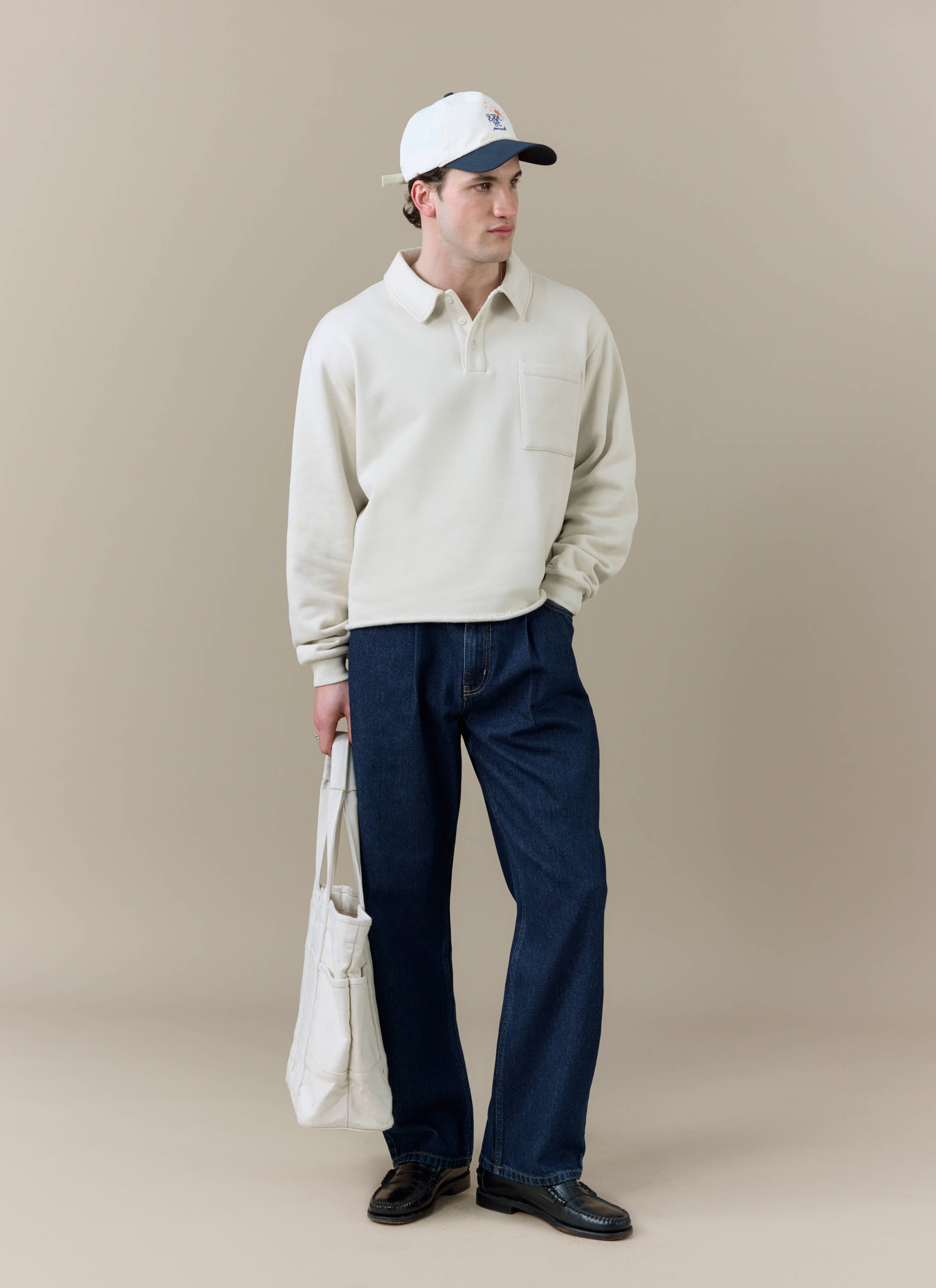 Full-length shot of a model wearing the Percival Dudley Boxy Sweatshirt in cream, with dark denim, a cap and a tote bag.