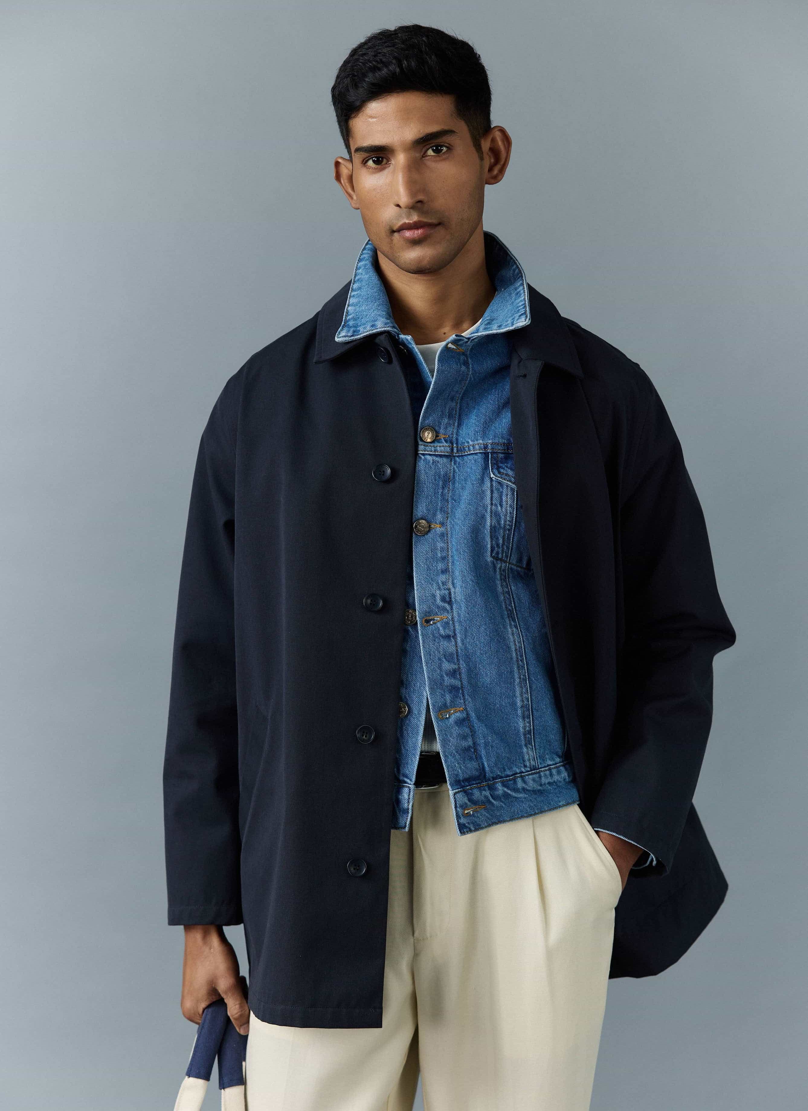 A model wears the navy Sherlock Trench Coat from Percival, layered over a denim jacket and cream trousers.