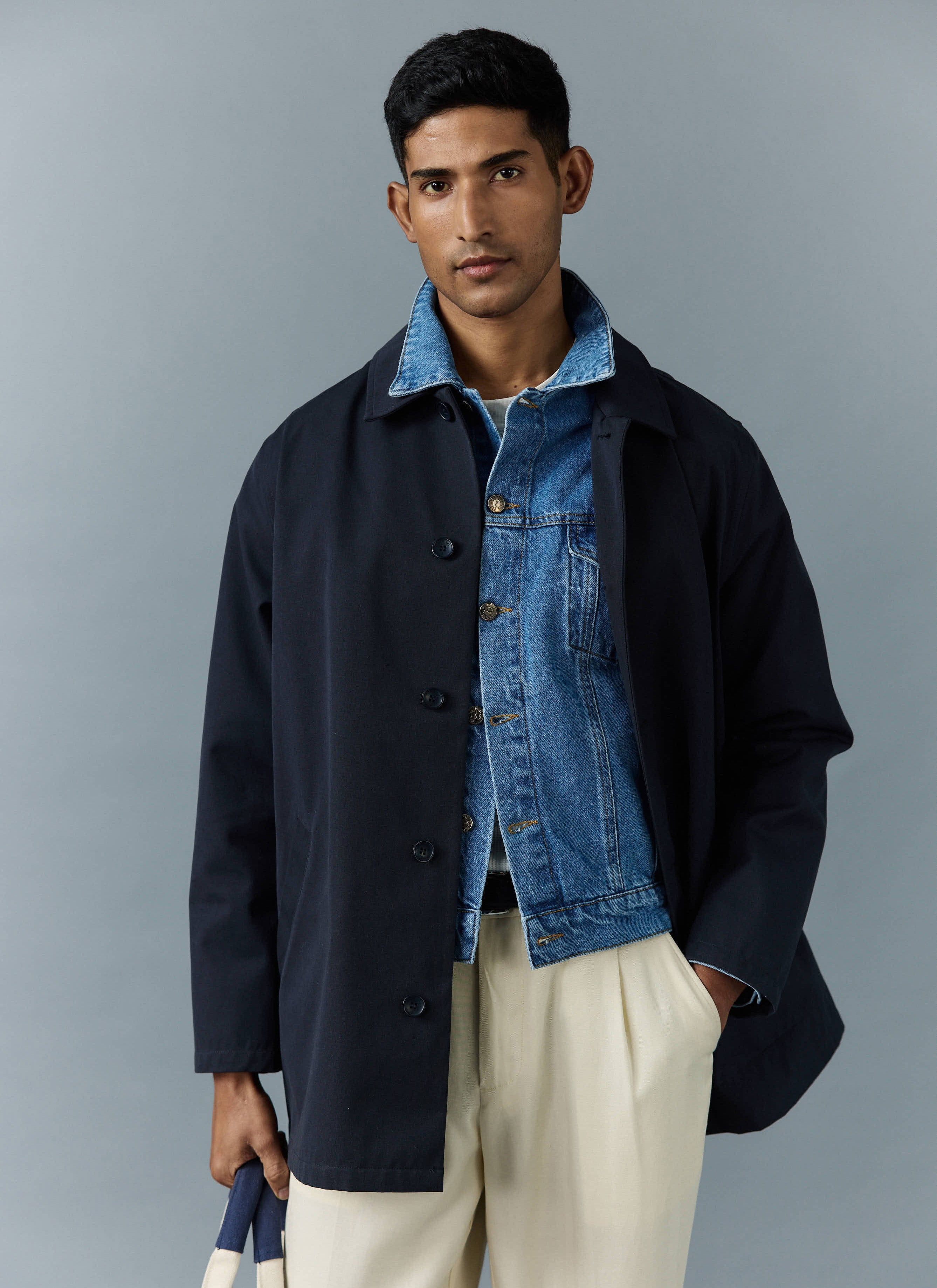 A model wears the navy Sherlock Trench Coat from Percival, layered over a denim jacket and cream trousers.