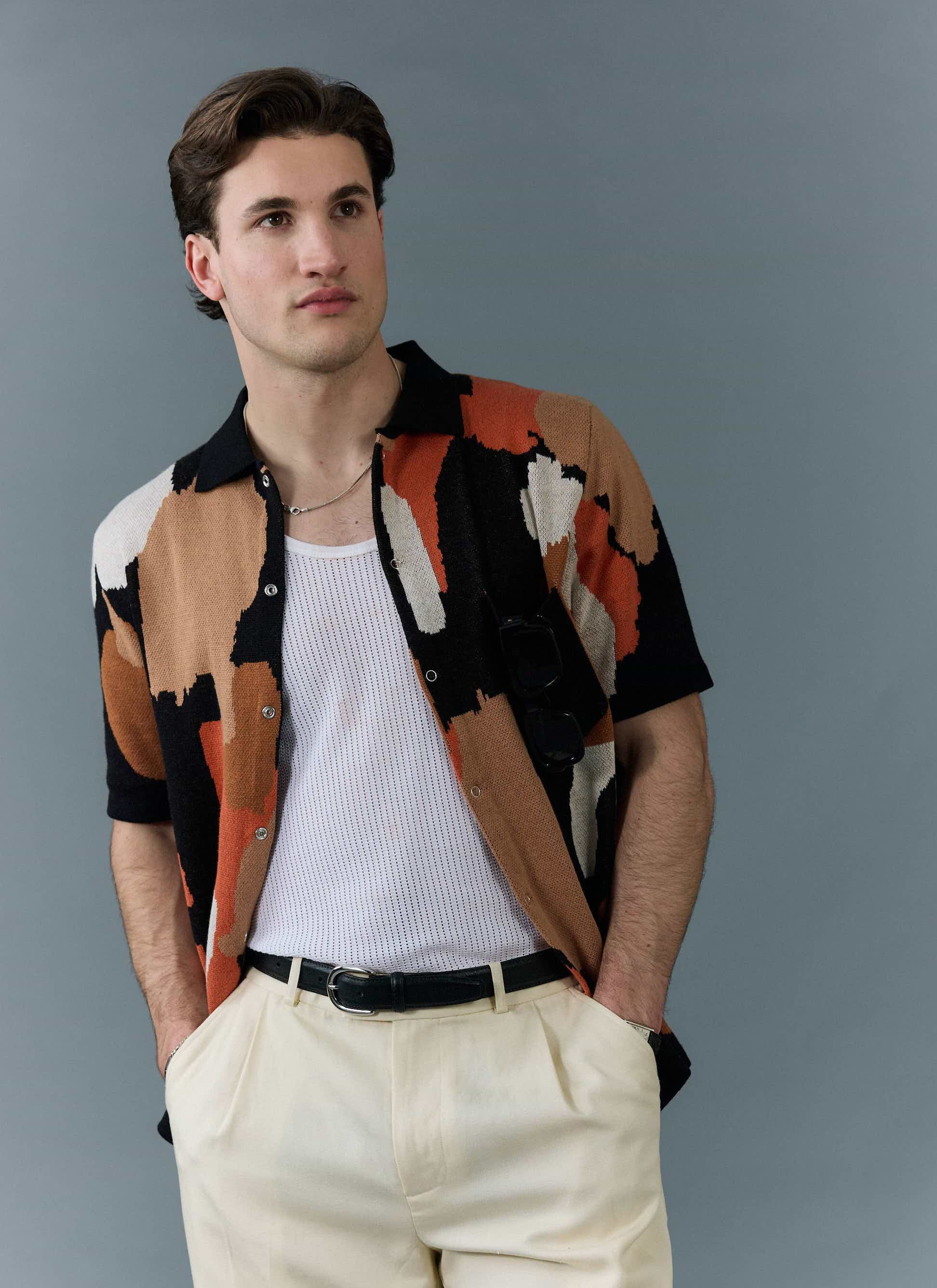 A male model wears the Percival Impressionist Jacquard Shirt in black, styled open over a white vest with cream trousers.